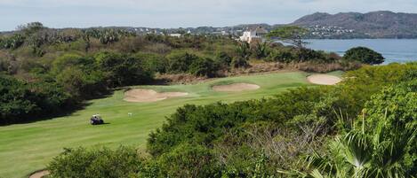 A golf course with bunkers, a water hazard, and a distant view of a coastal town.