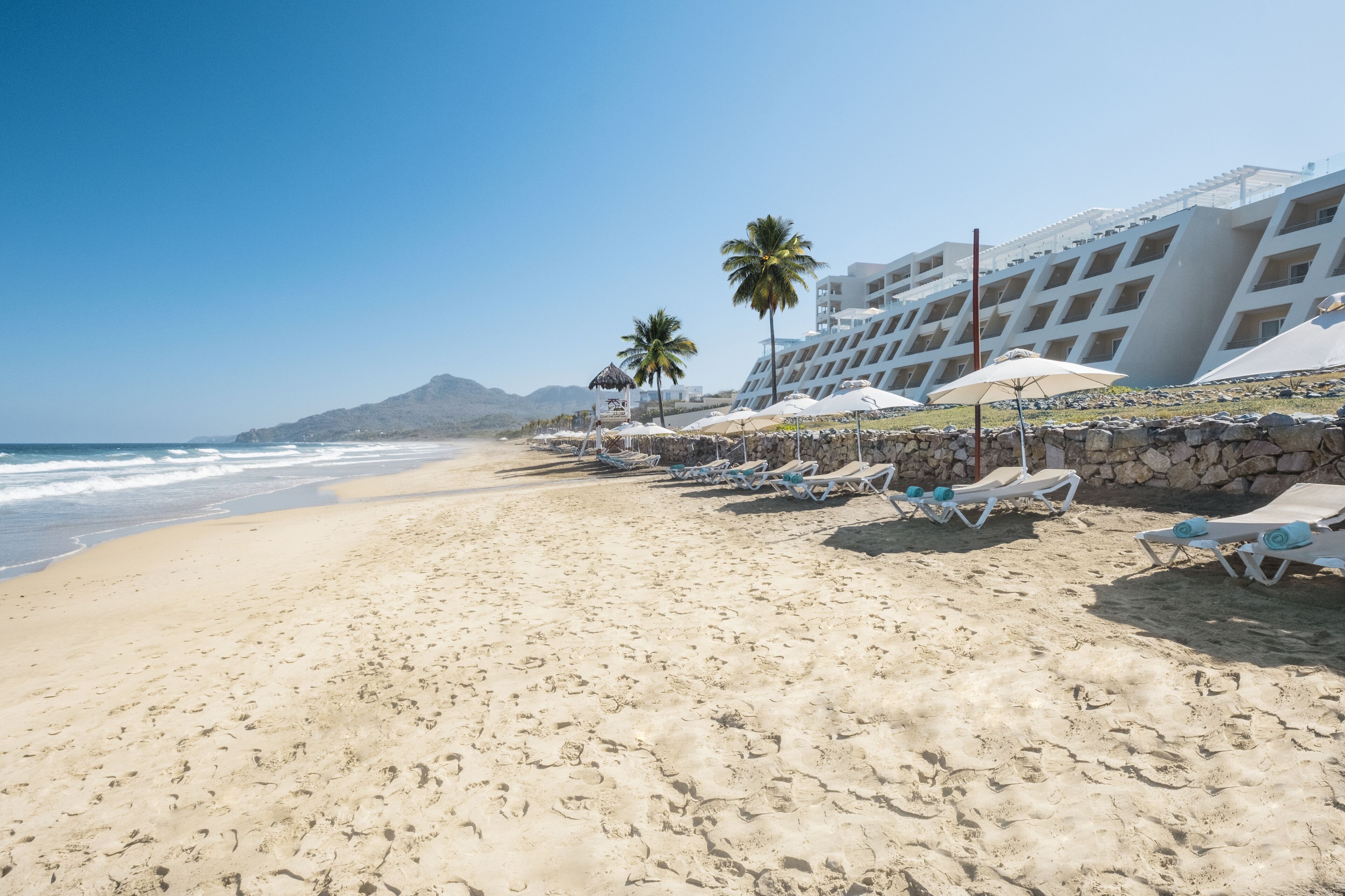 Een strand met ligbedden, parasols en een hotelgebouw met meerdere verdiepingen en balkons.