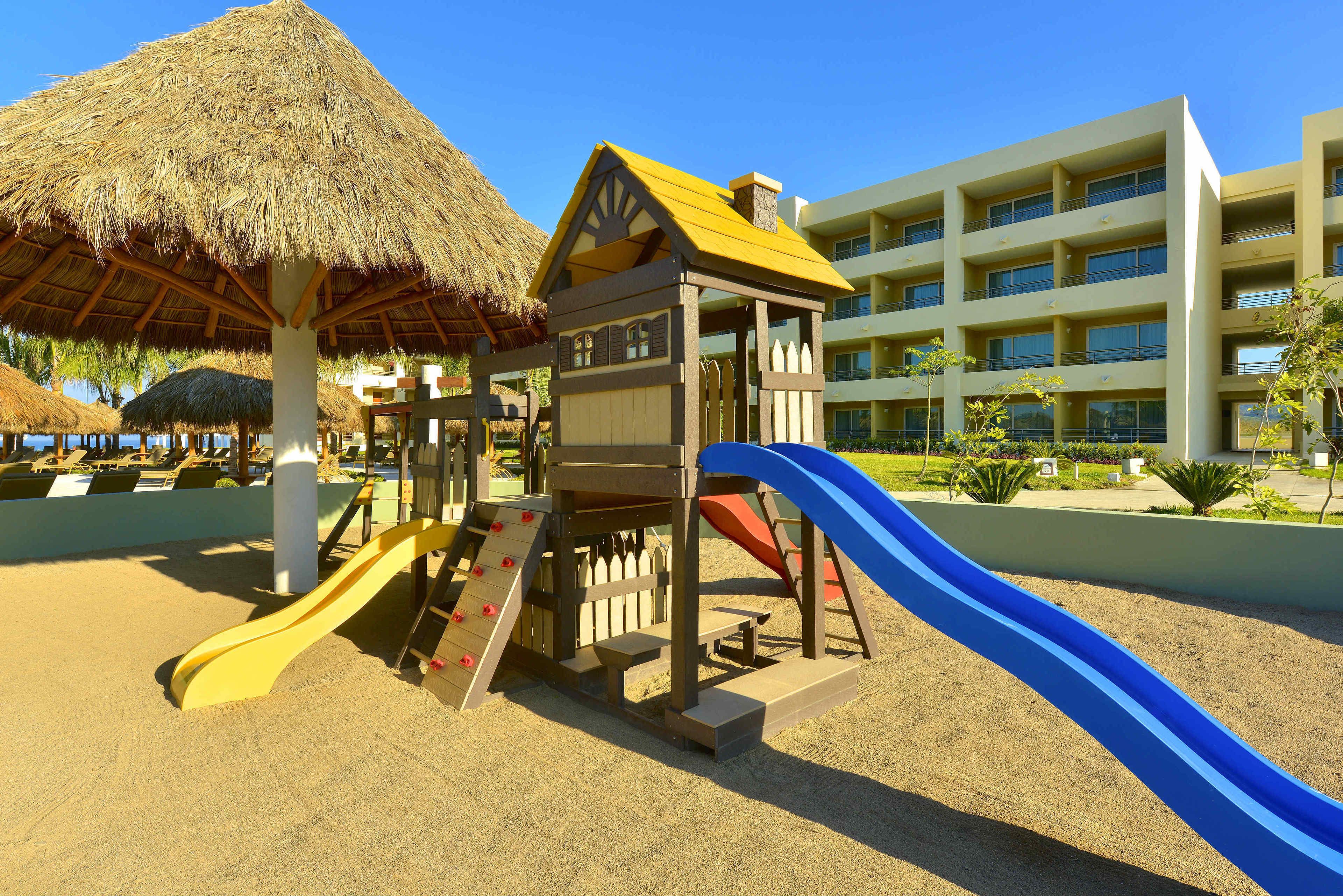 A playground with slides and a thatched-roof structure, adjacent to a multi-story building.