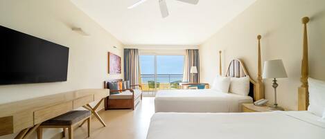 A hotel room with a bed, a desk, a TV, a ceiling fan, and a view of the ocean.