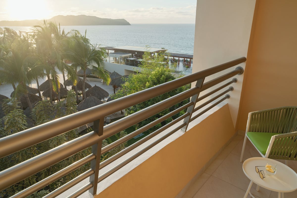 A balcony with a view of the sea, a table with a chair, and a thatched-roof structure.