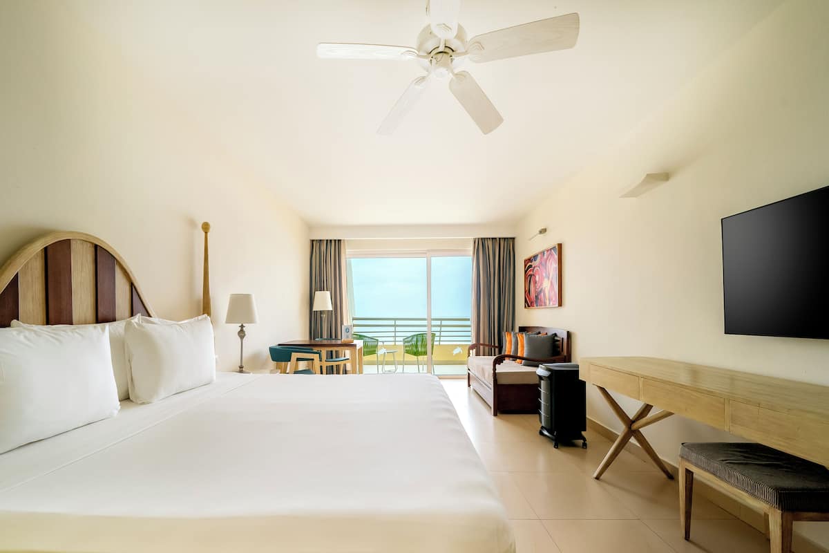 A hotel room with a large bed, a desk with a chair, a television, and a balcony with a view.