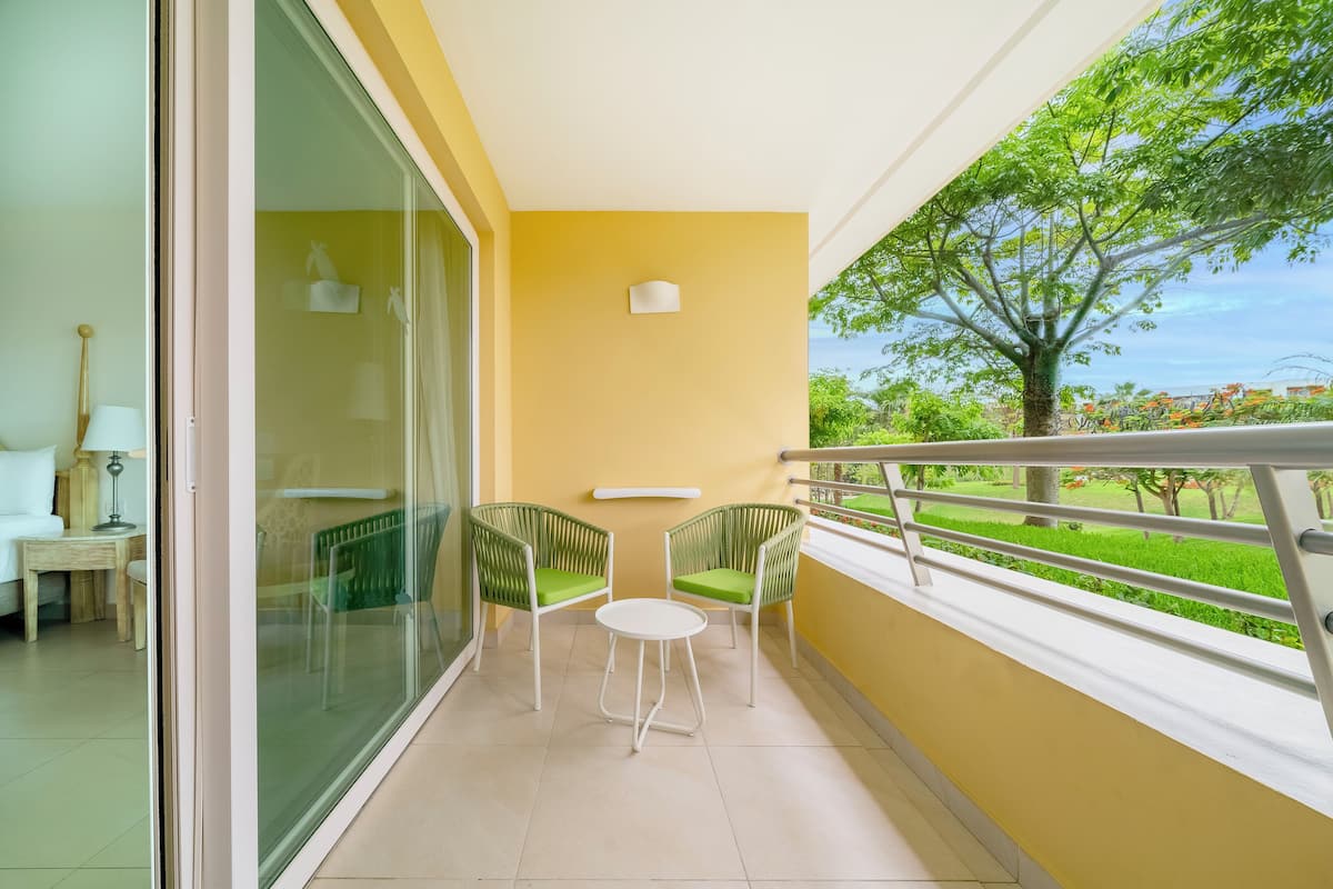 A balcony with a glass door, two wicker chairs with green cushions, a small white table, and a view of greenery and buildings.