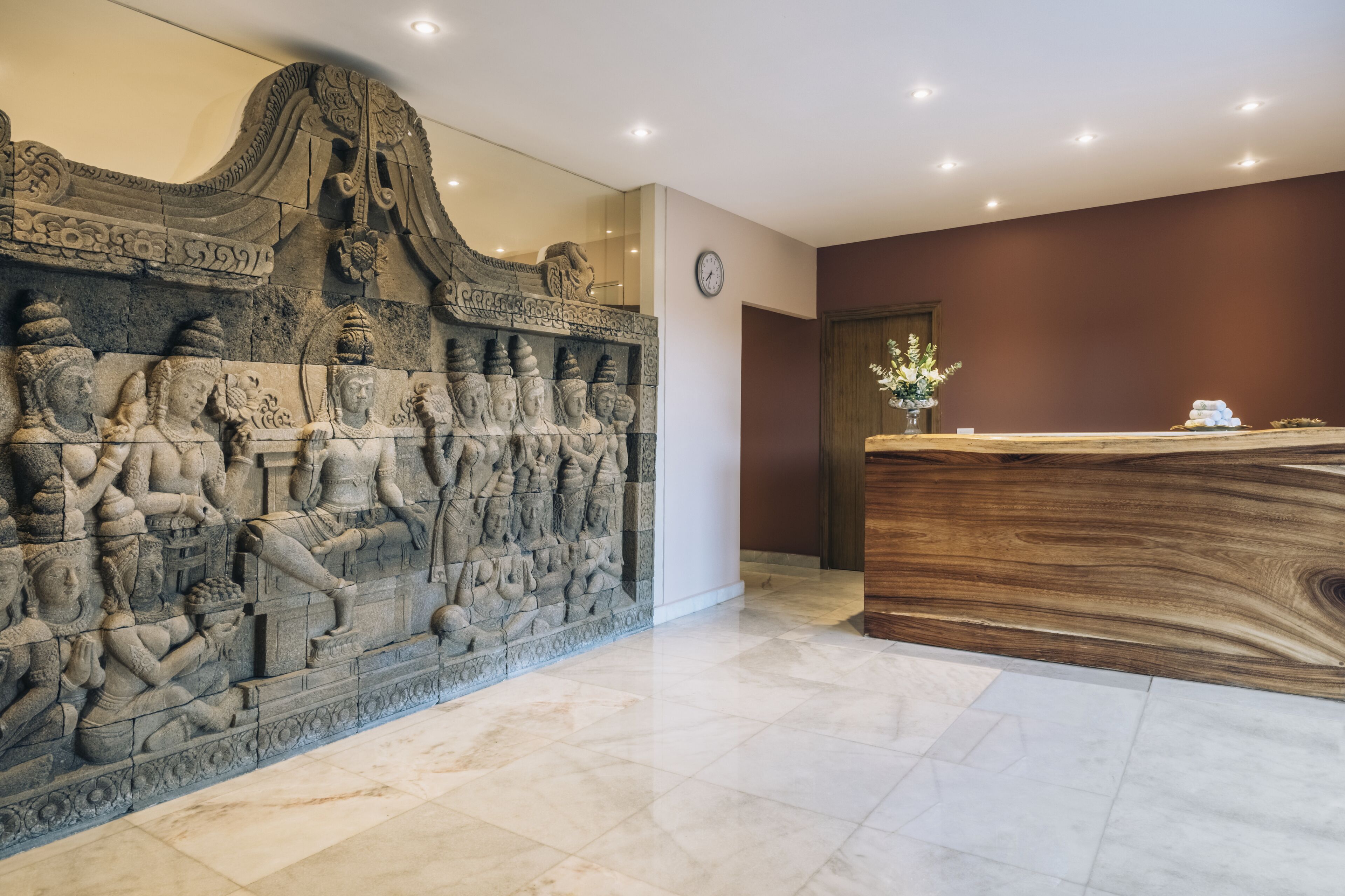 A reception area with a stone wall featuring intricate carvings, a wooden reception desk, and a clock on the wall.