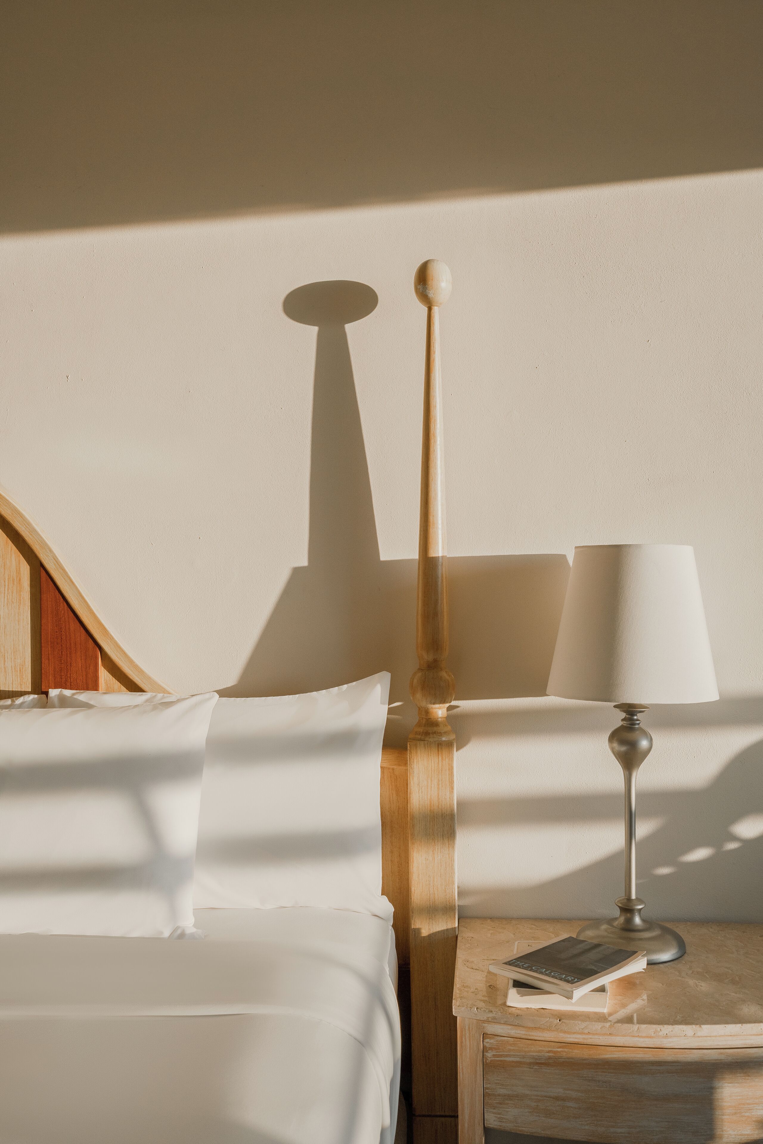 Un lit bien fait, avec du linge de lit blanc, une table de chevet en bois sur laquelle se trouve une lampe, et un livre posé sur la table de chevet.