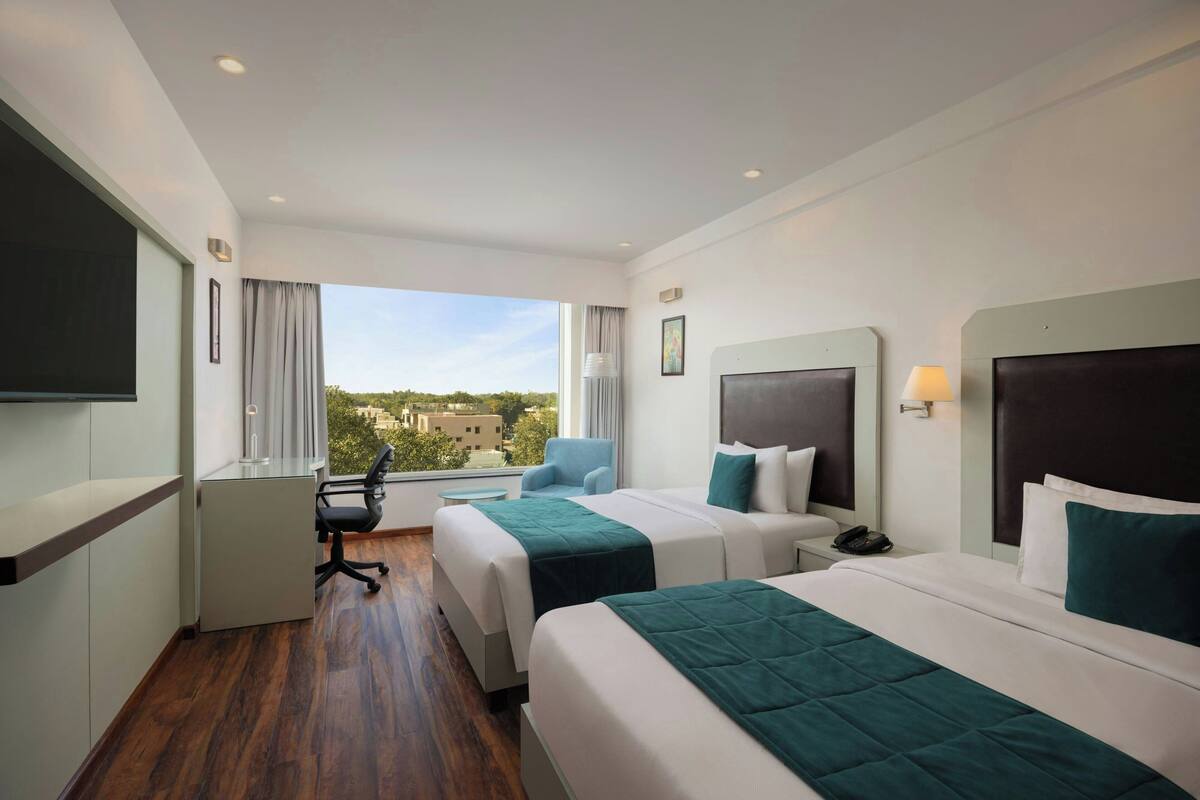 Ein Hotelzimmer mit zwei Betten, einem Schreibtisch, einem Stuhl, einem Fernseher und einem Fenster mit Blick auf die Stadt.