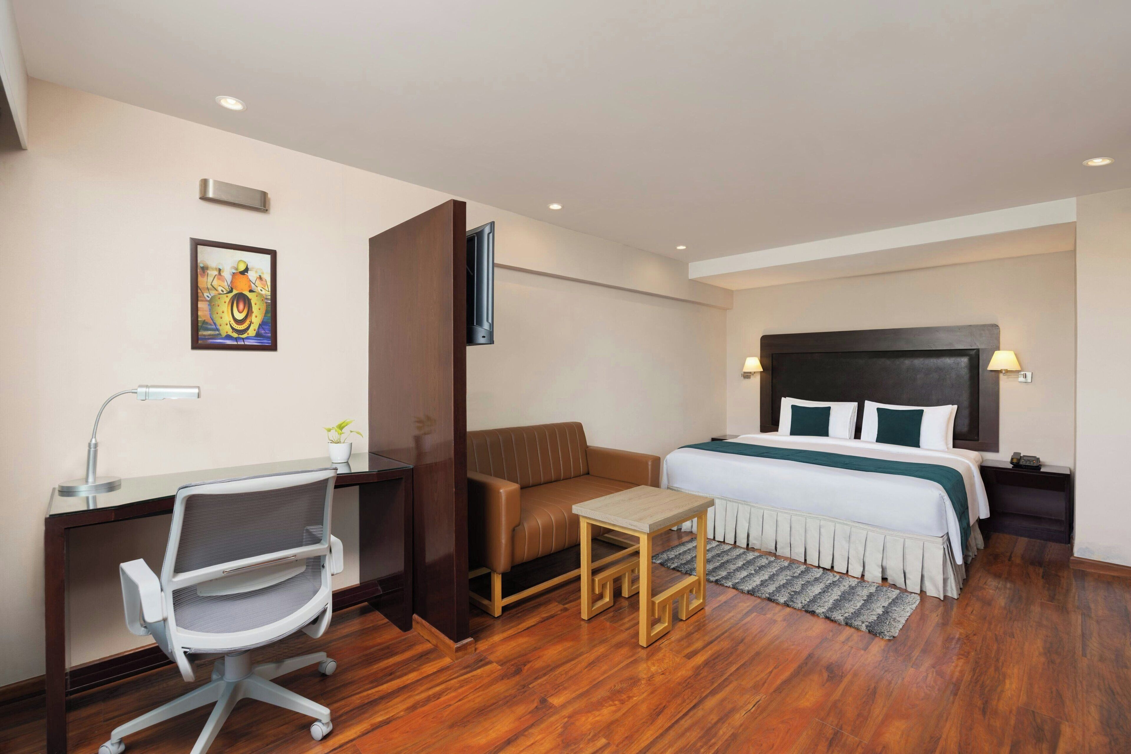 A hotel room with a bed, a desk with a chair, a small table, a sofa, a painting on the wall, and a television.