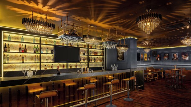 A bar area with a long counter, multiple bar stools, and a variety of bottles and glasses on shelves.