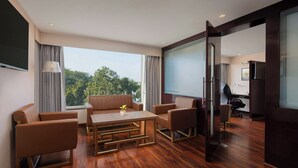 A modern hotel room with a wooden floor, a sofa set, a dining table, and a large window offering a view of trees.