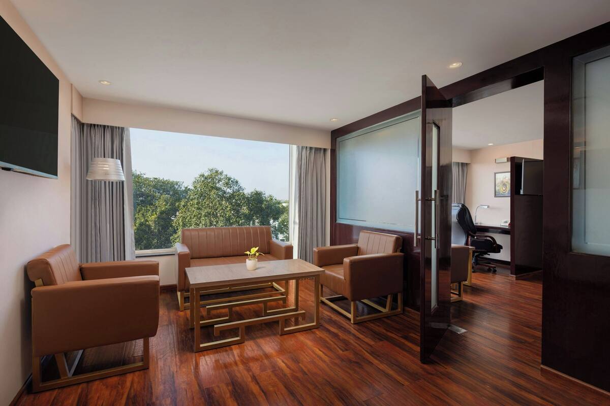 Ein modernes Hotelzimmer mit Holzboden, einer Sofagarnitur, einem Esstisch und einem großen Fenster mit Blick auf Bäume.