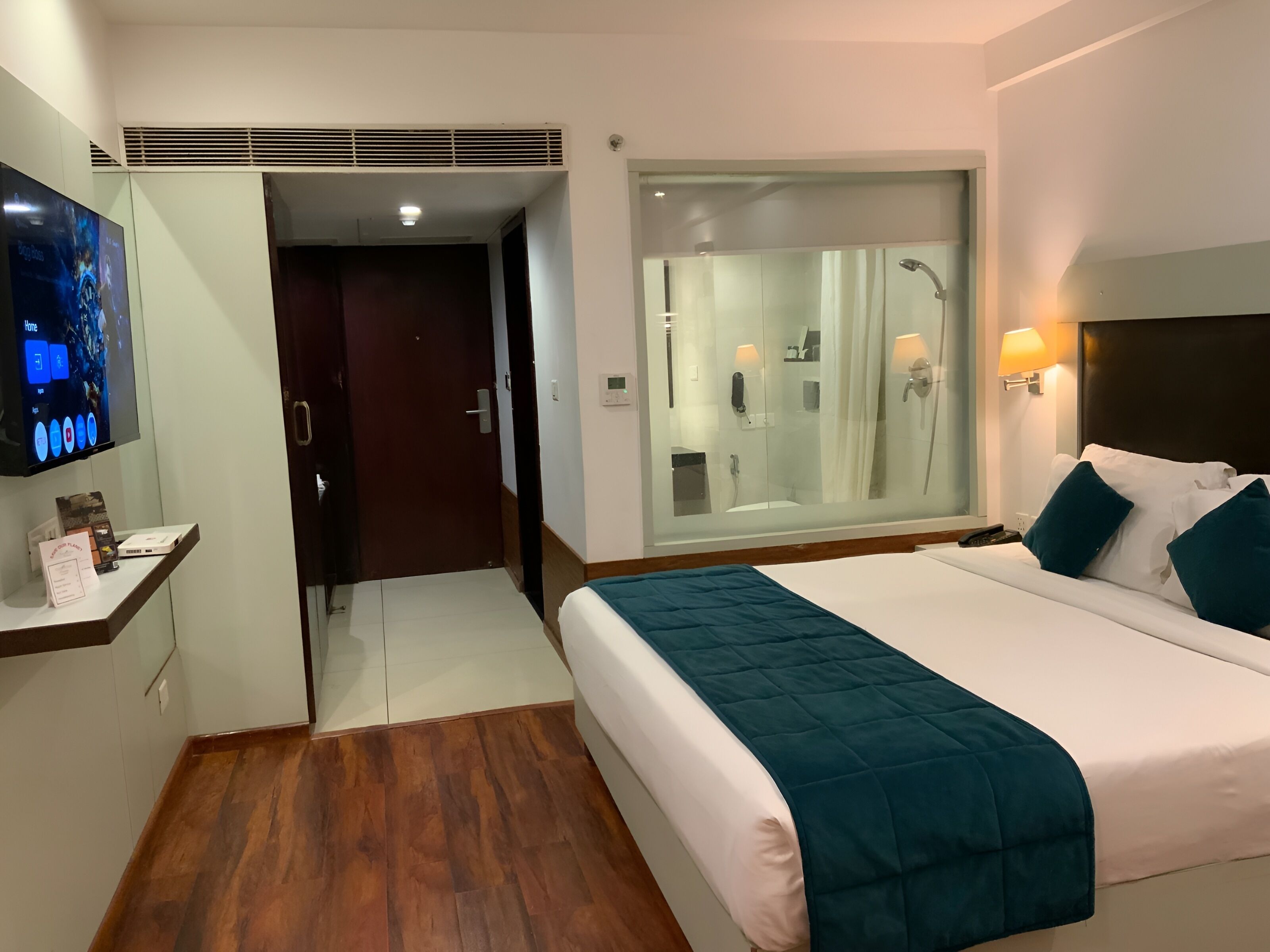 Habitación de hotel con cama, televisor, baño con cabina de ducha de vidrio y suelo de madera.