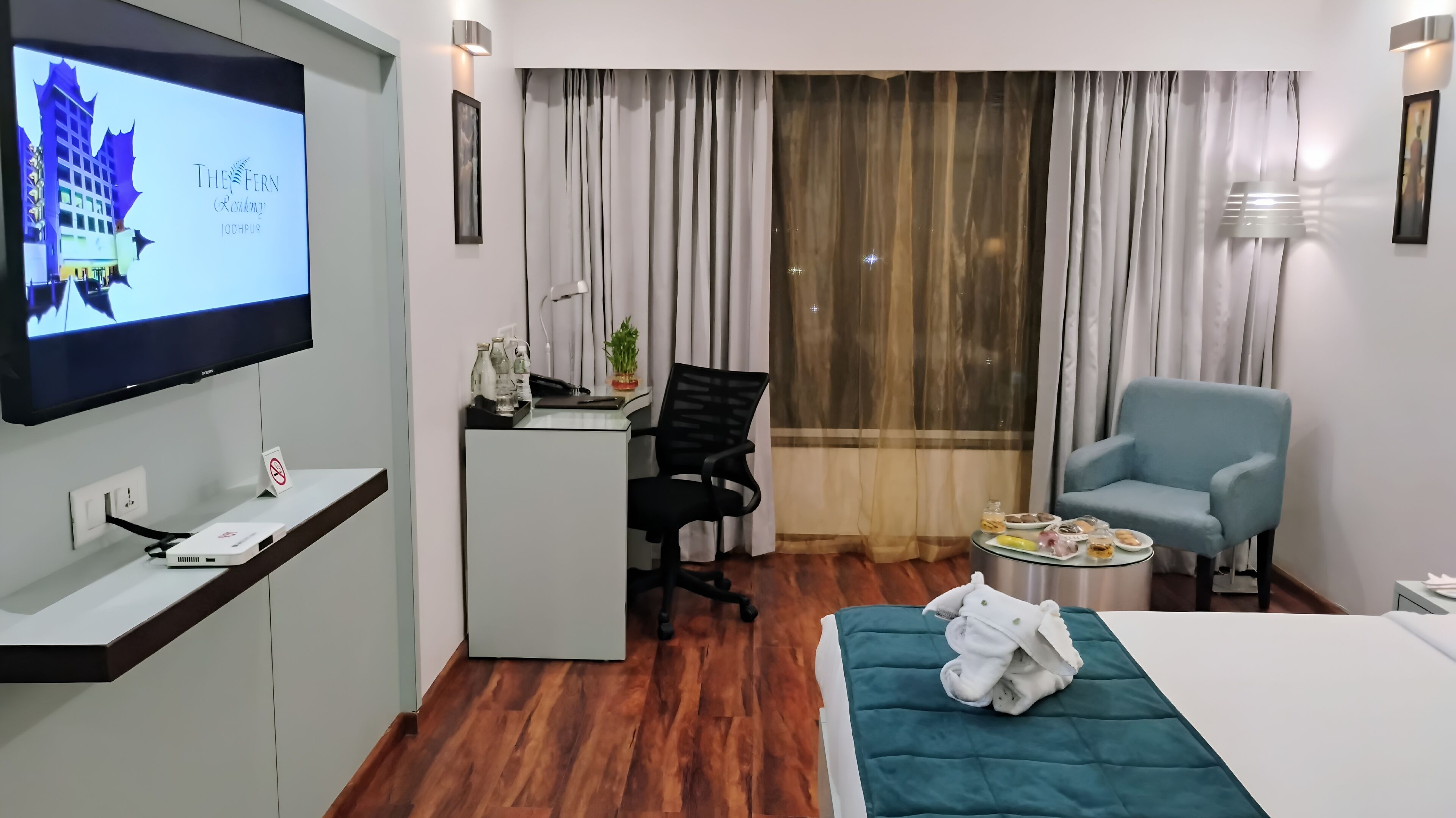 A hotel room with a bed, a desk with a computer, a chair, a TV, and a window with curtains.