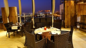 An outdoor dining area with wicker furniture and a table set for two, overlooking a cityscape at dusk.