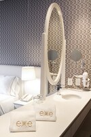 Un baño de hotel con papel tapiz estampado, un espejo ovalado grande, un lavamanos blanco y dos toallas con la palabra "exxe" bordada.