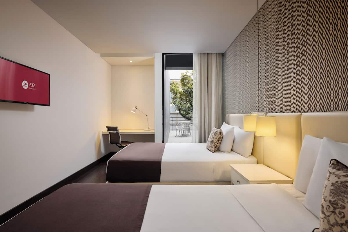 Habitación de hotel con dos camas, un escritorio, televisor y vista al exterior.