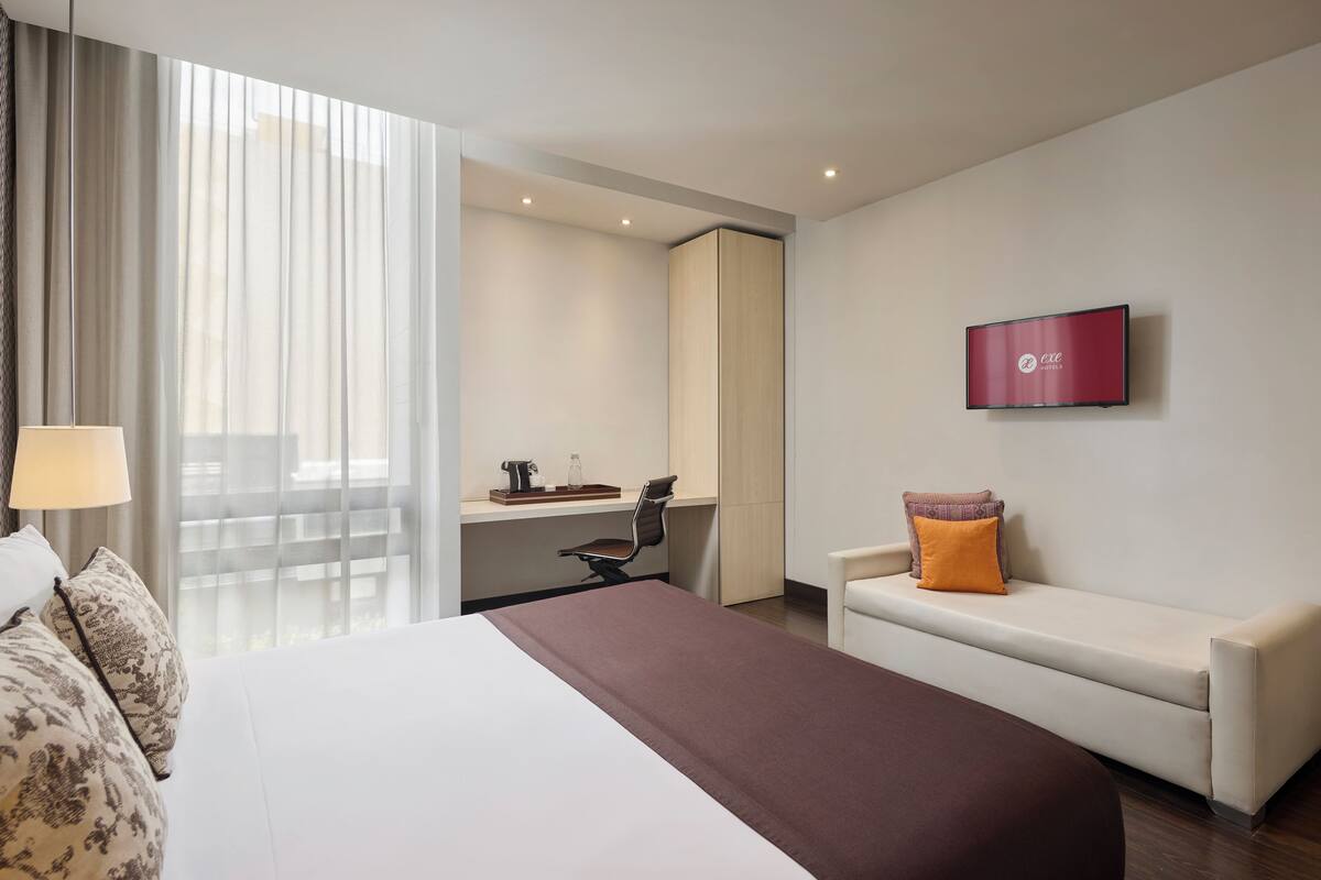 Habitación de hotel con cama, escritorio, silla, televisor y ventana con cortinas.