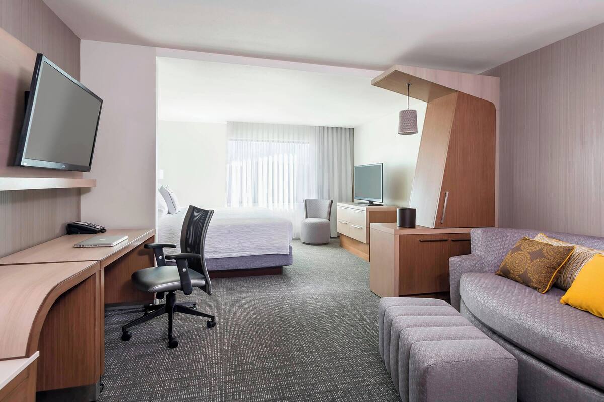 A hotel room with a bed, a desk with a computer, a TV, a sofa, and a TV on a stand.