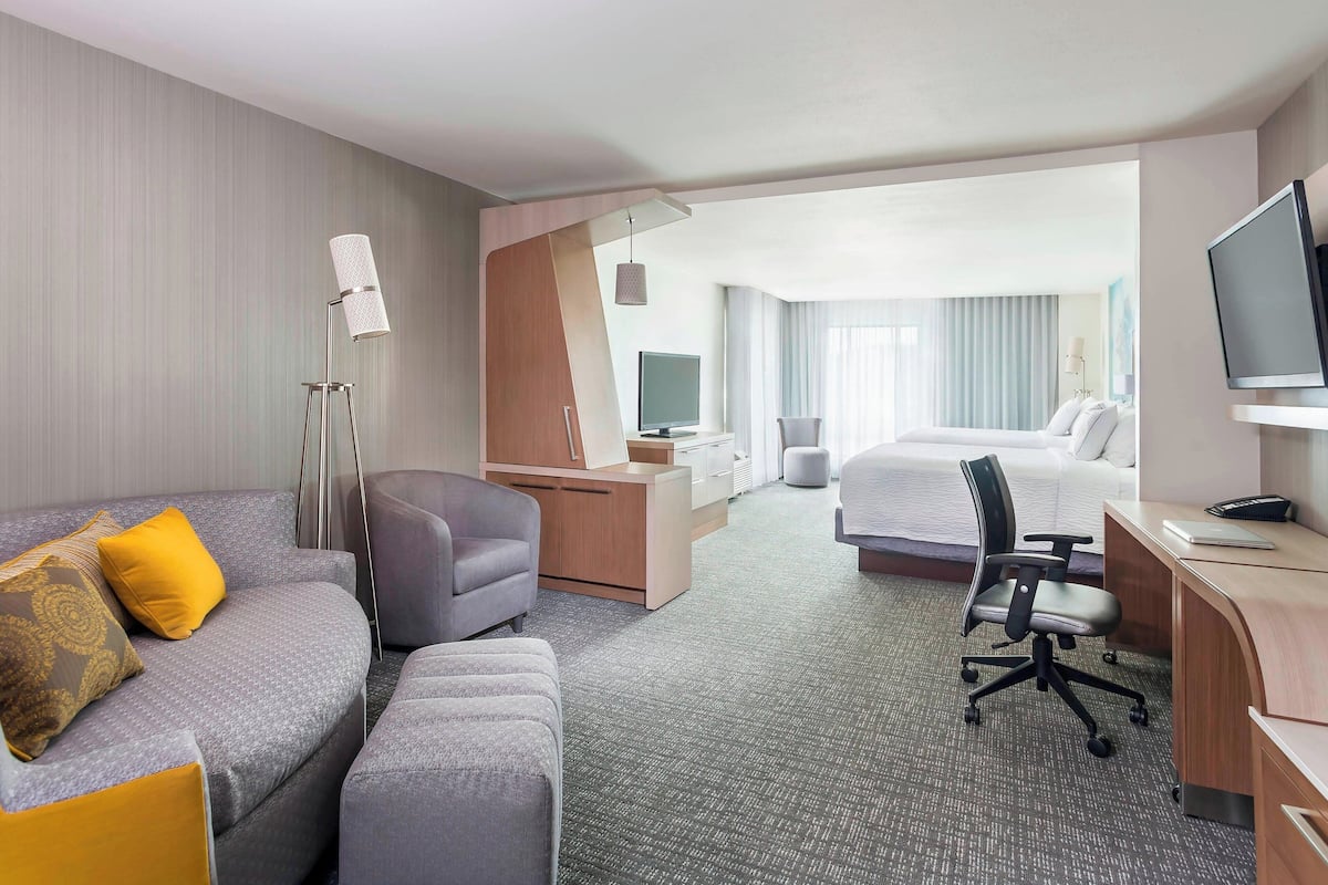 A hotel room with a grey sofa, a desk, a bed, a TV, and a desk lamp.