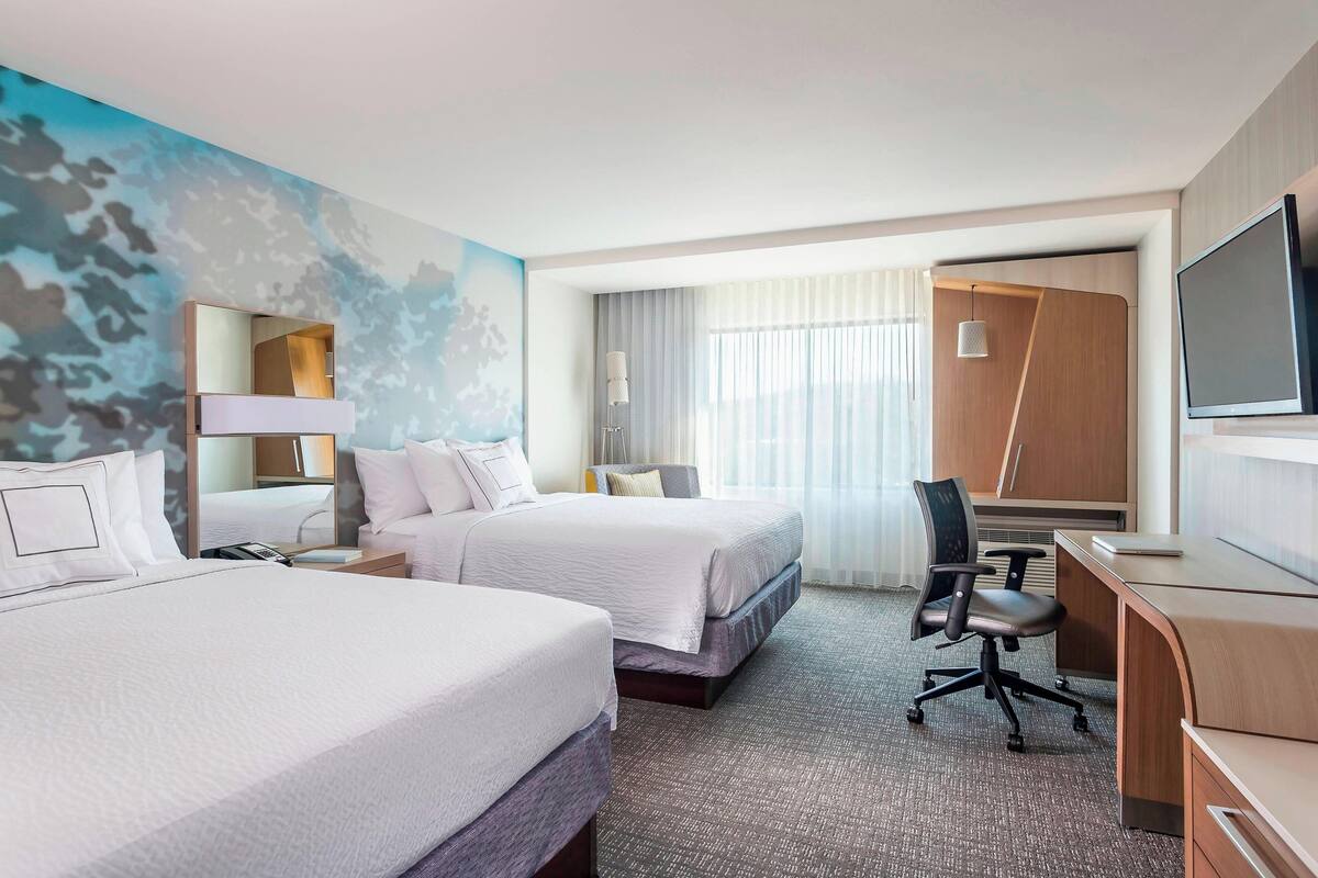 A hotel room with two beds, a desk, a TV, and a large window with curtains.