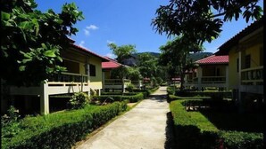 A pathway leading to a row of yellow buildings with red roofs, surrounded by well-maintained greenery and a clear blue sky. - New Sunrise Village Bang Po Beach (Koh Samui)