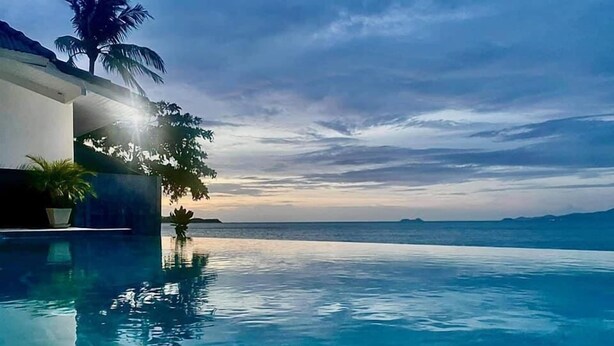 An infinity pool with clear blue water, a palm tree, and a view of the ocean at sunset. - The Cosy Beach Resort (Koh Samui)
