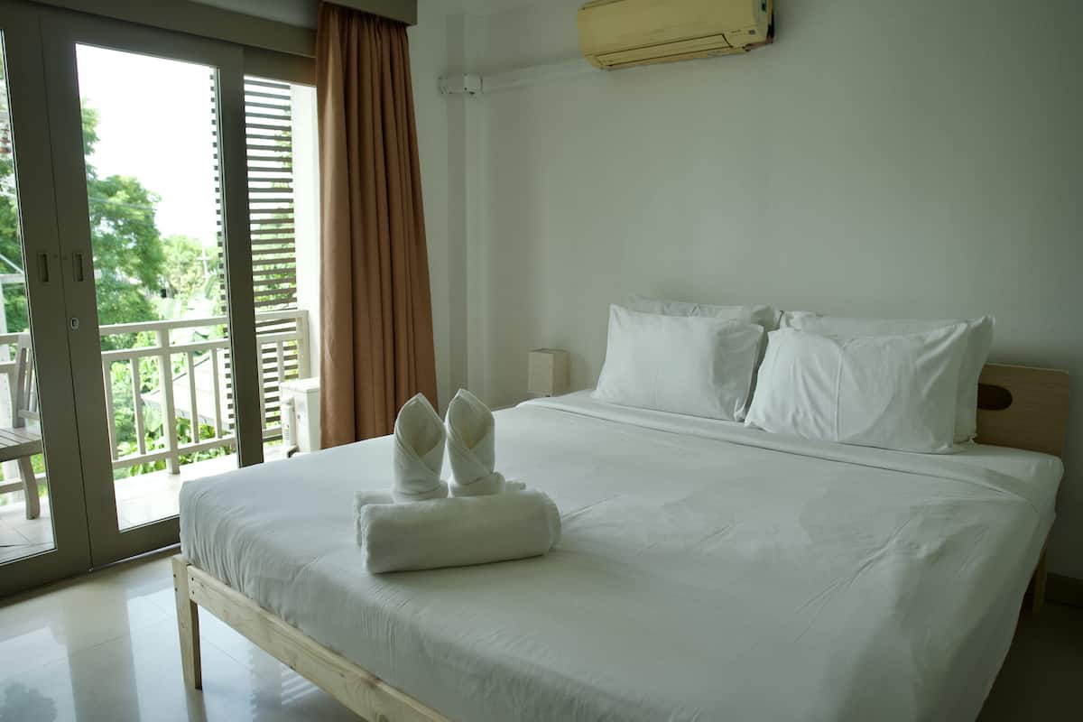 A neatly made bed with white linens and towels, a wooden headboard, and a view of greenery through a sliding glass door.