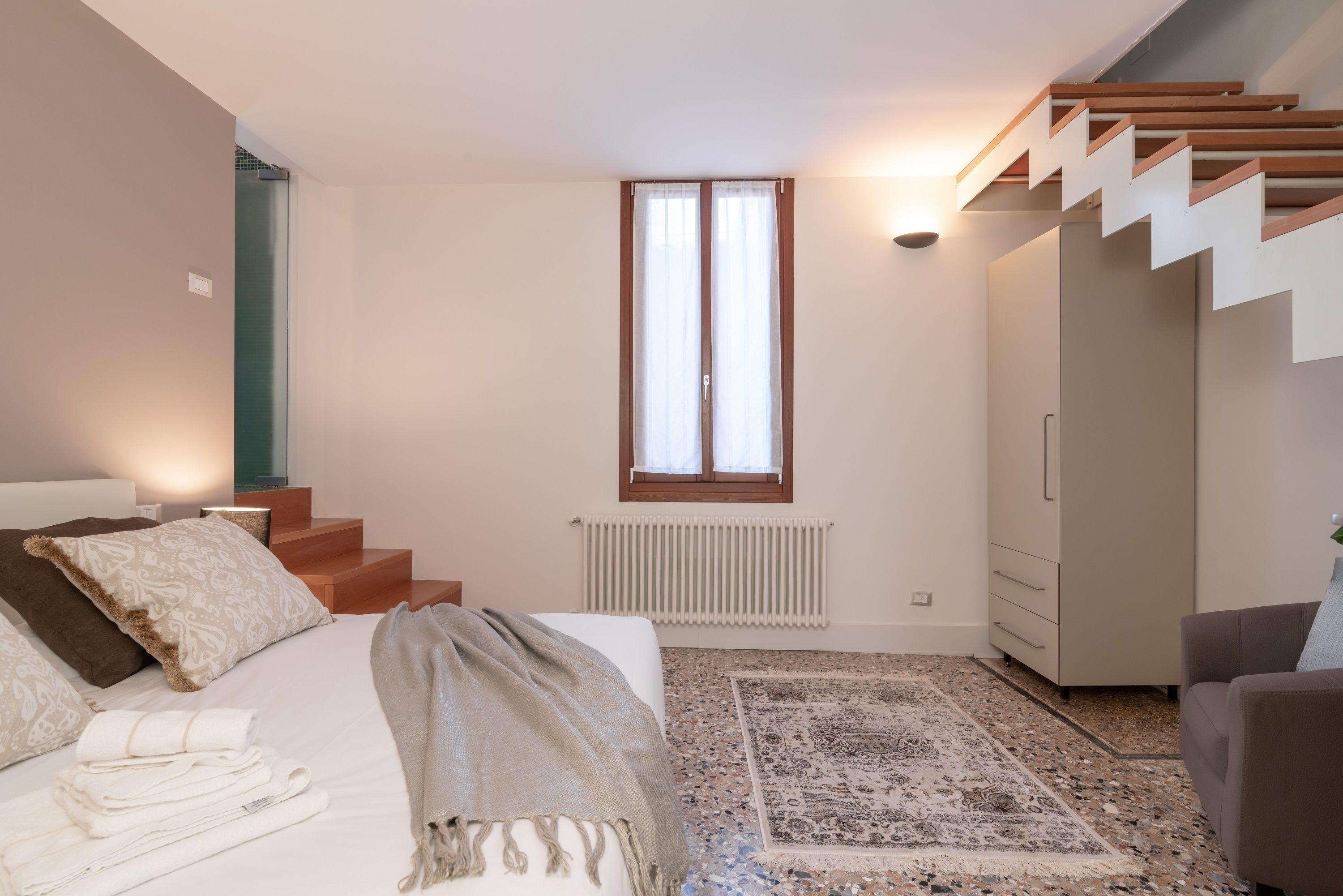 Een slaapkamer met een bed, een raam, een radiator, een trap en een kledingkast.