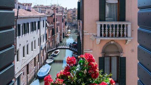 A narrow canal with boats, a pink building with shutters, and a balcony with flowers.