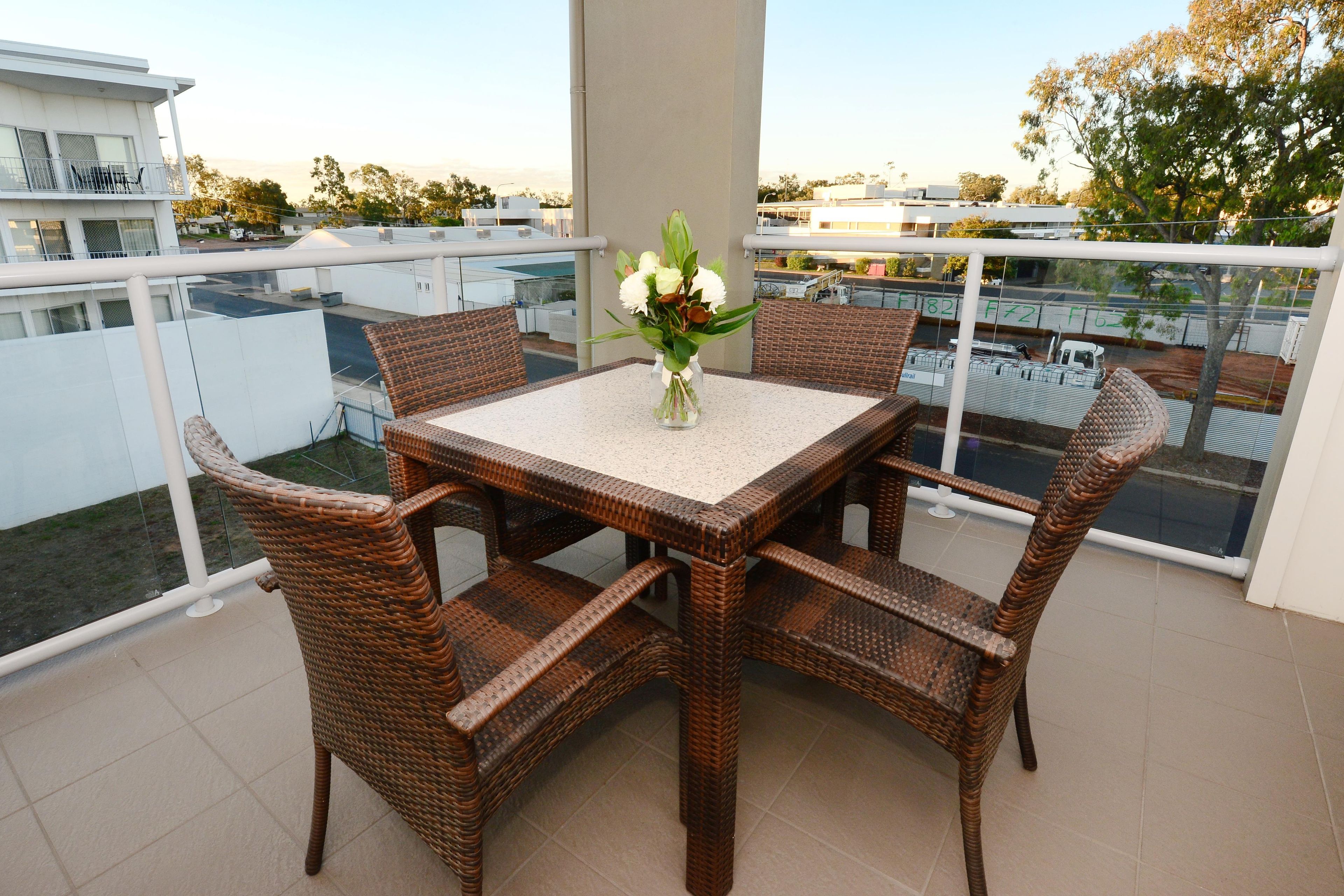 A balcony with a table and four wicker chairs, a vase with flowers, and a view of the cityscape.