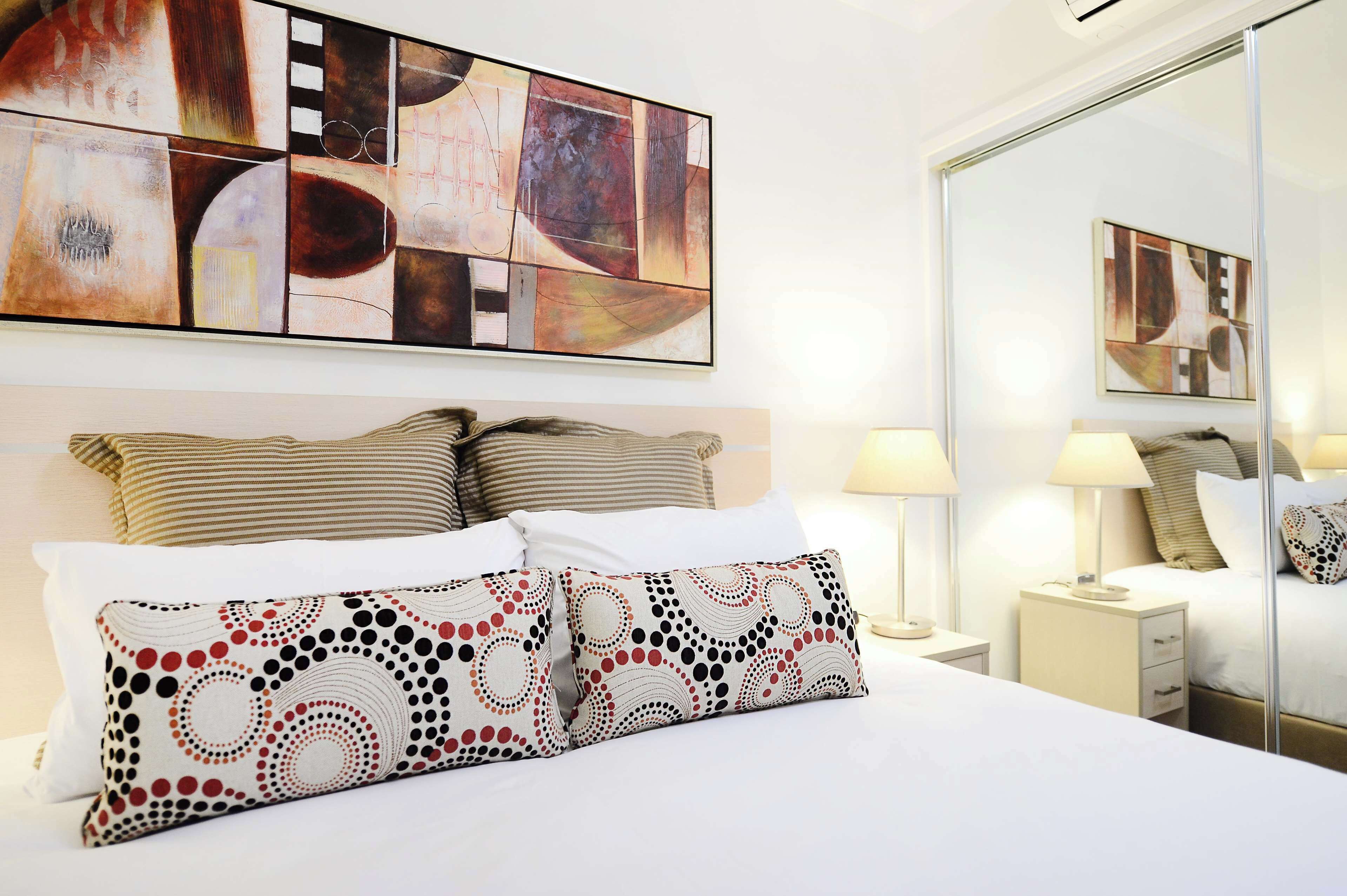 A hotel room with a large abstract painting above the bed, a bed with striped and patterned pillows, bedside lamps, and a mirror reflecting the room.