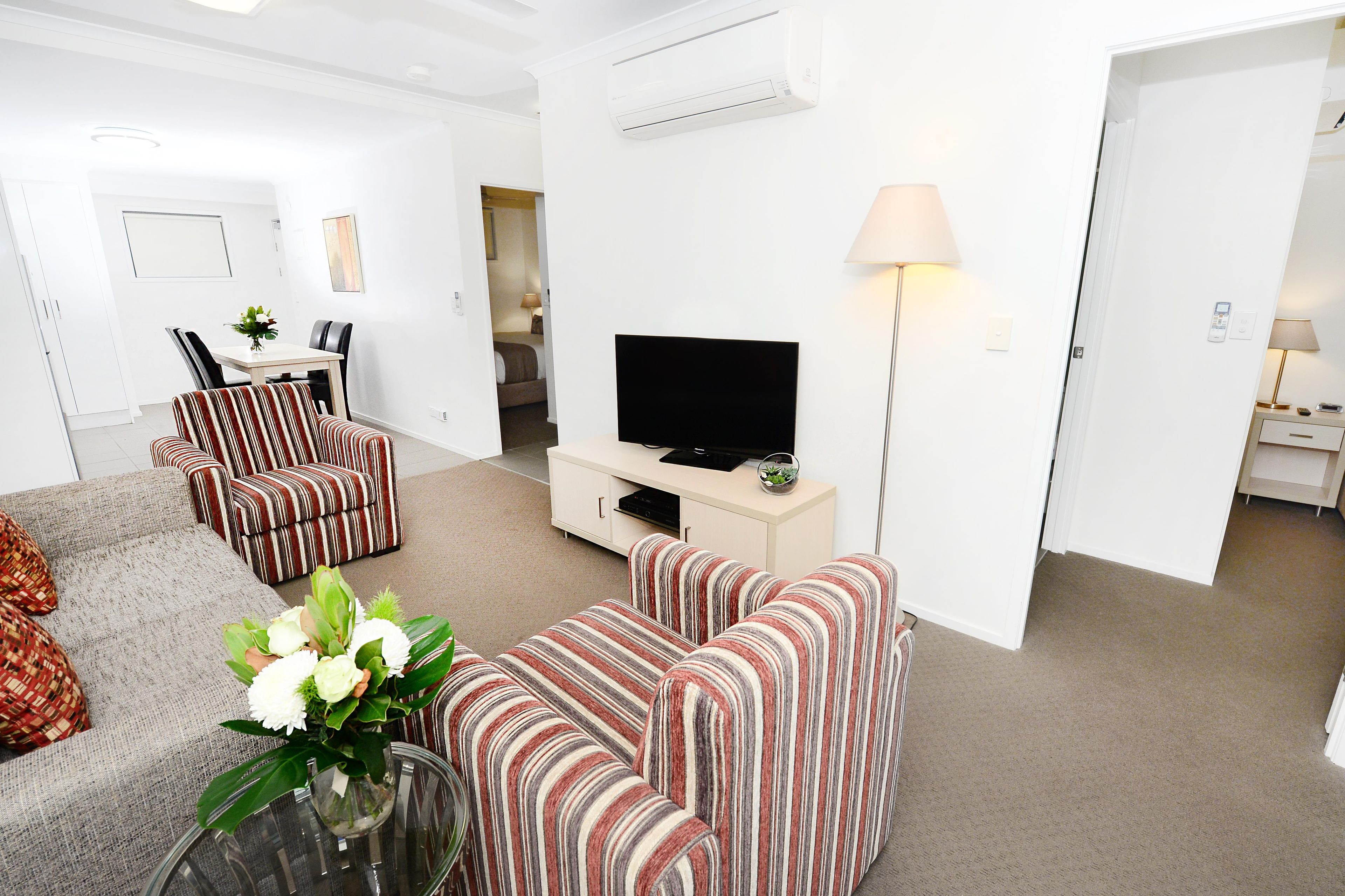 En moderne stue med stribede sofaer, et fladskærms-tv, et spiseområde med bord og stole, og en gang der fører til et soveværelse.