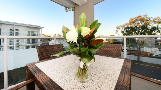 Un balcón con mobiliario de mimbre, un florero de cristal con flores y una vista despejada del paisaje urbano.