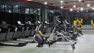 A row of stationary bikes and treadmills in a gym with large windows and a concrete floor.