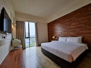 Ein modernes Hotelzimmer mit einem großen Bett, einem hölzernen Kopfteil, einem an der Wand montierten Fernseher, einem Schreibtisch mit Stuhl und Blick auf die Stadt durch das Fenster.