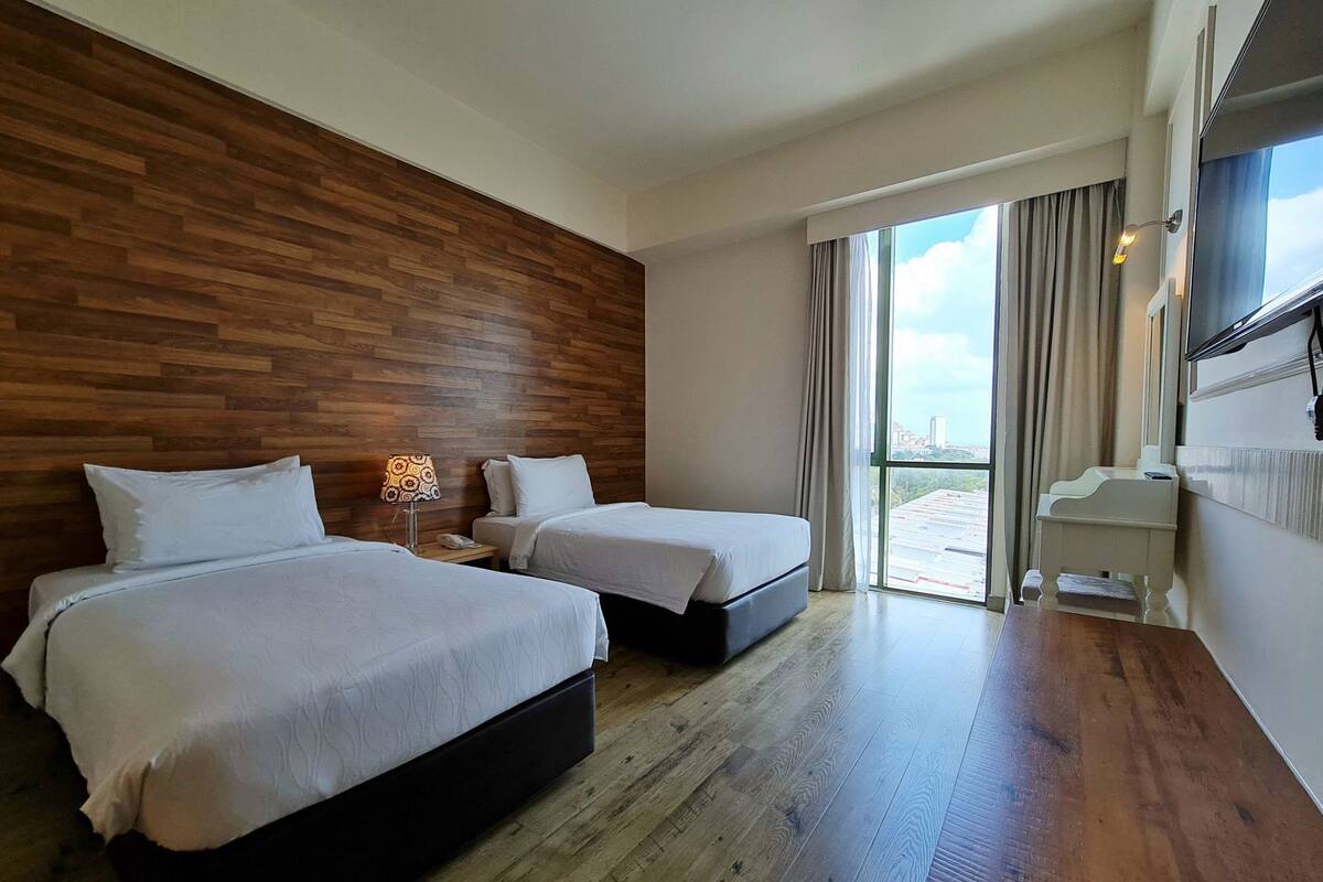 A hotel room with two beds, a wooden accent wall, a large window with a city view, and a small table with a lamp.