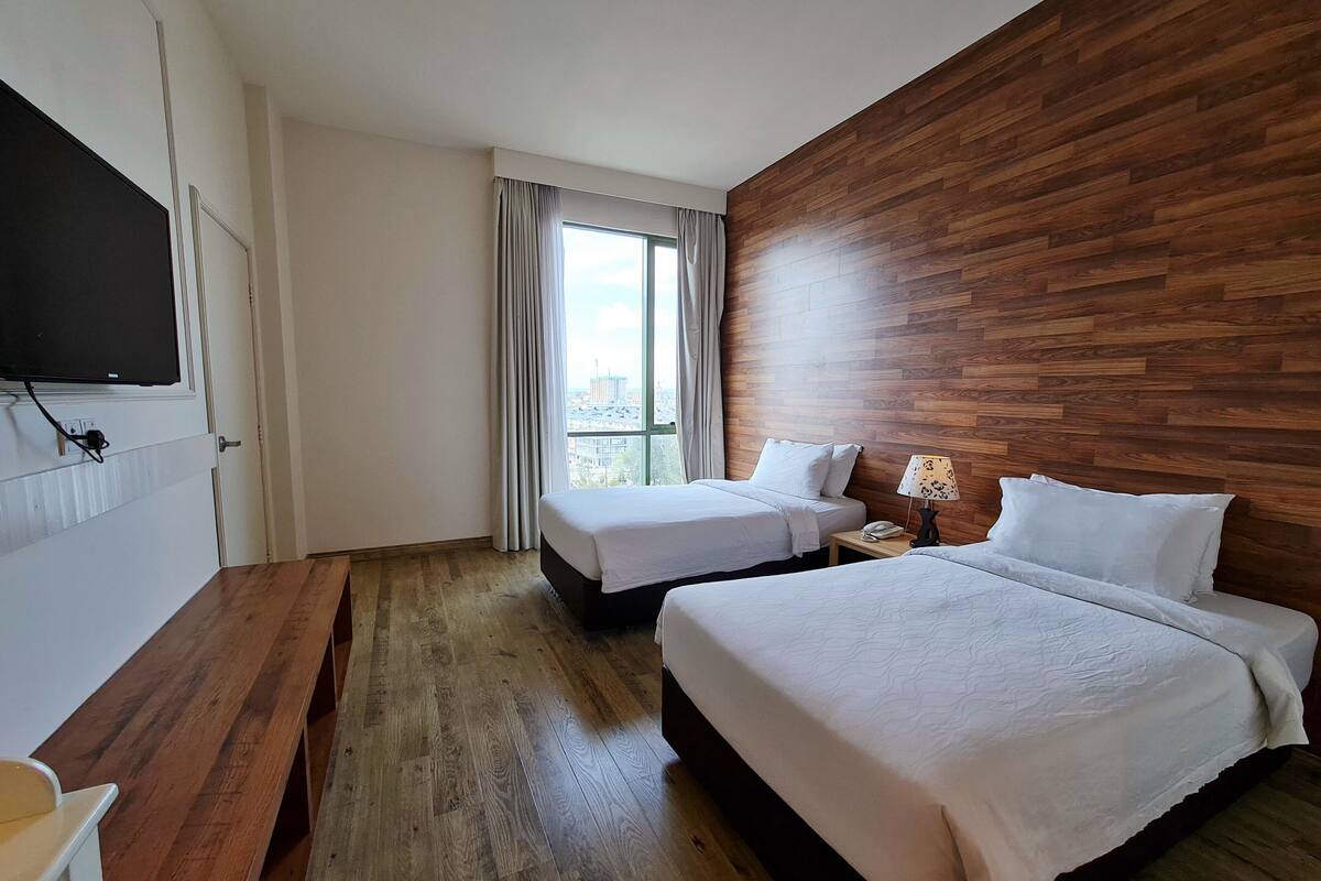 A hotel room with two beds, a wooden accent wall, a large window with curtains, a bedside table with a lamp, and a TV mounted on the wall.