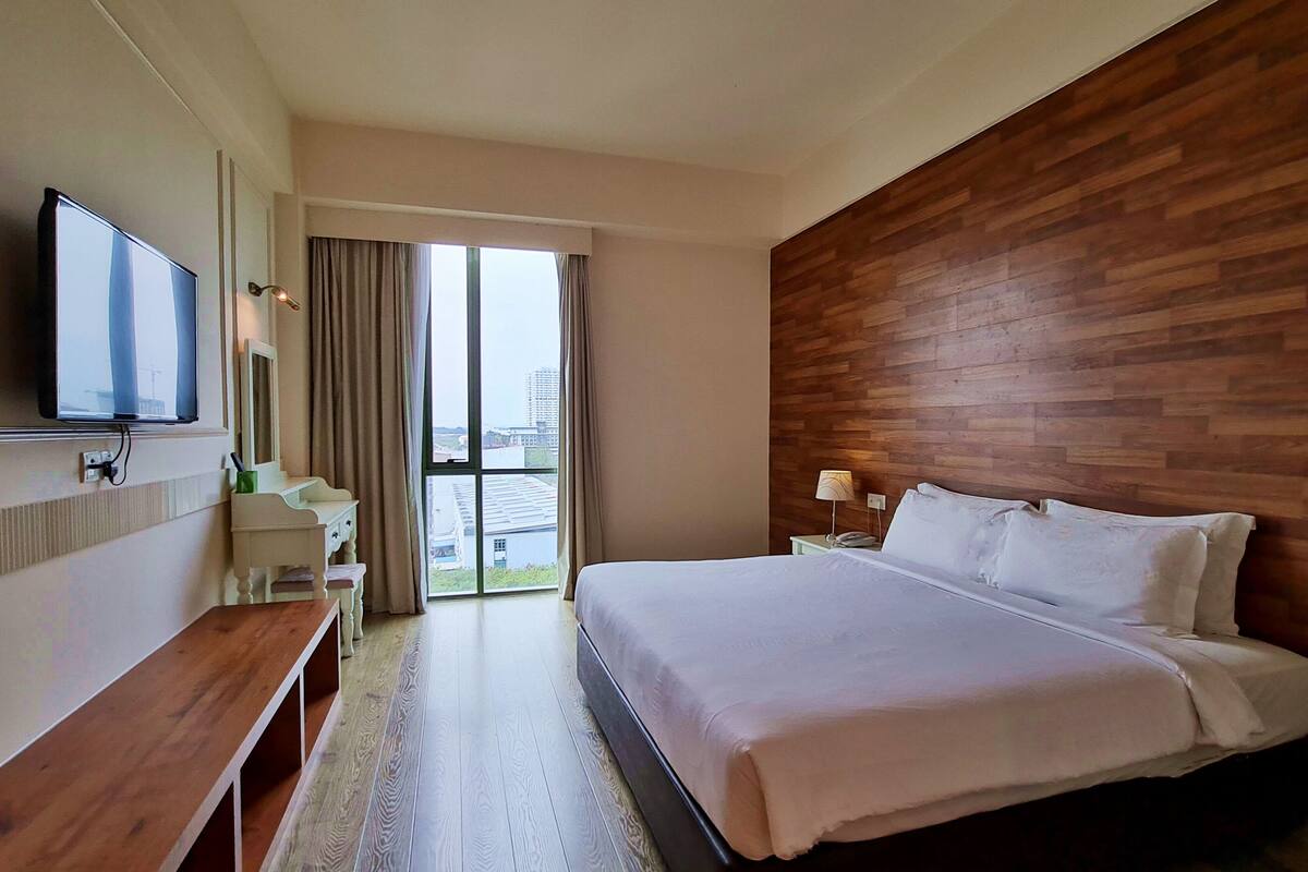 A hotel room with a large bed, a wooden accent wall, a television, a desk, and a window with curtains.
