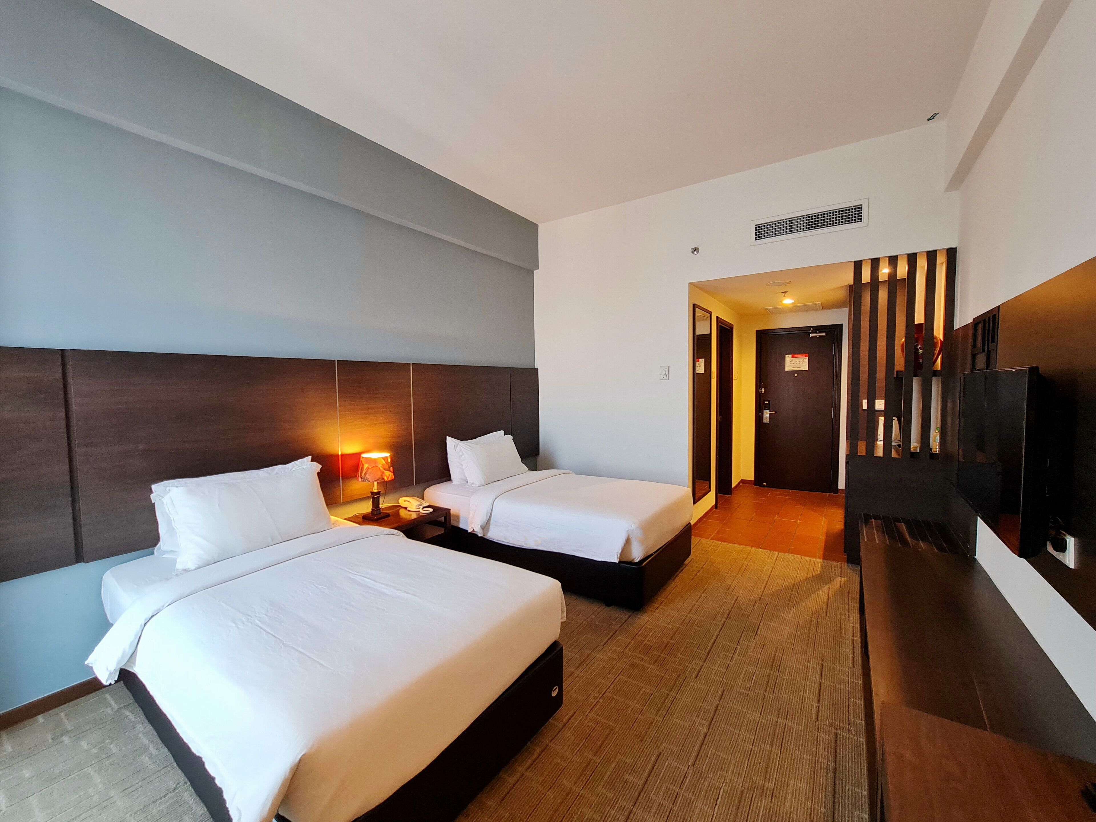 A hotel room with two beds, a TV, a desk with a lamp, and a door leading to another room.