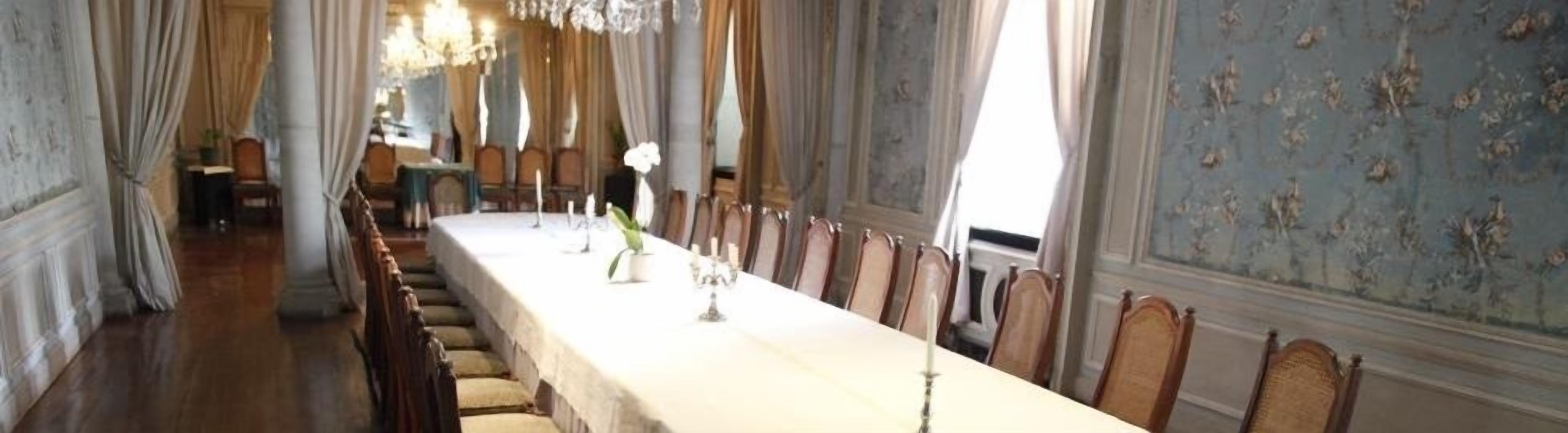 Une longue table à manger, entourée de chaises, dans une pièce à l’architecture classique et aux murs recouverts d’un papier peint à motifs floraux.
