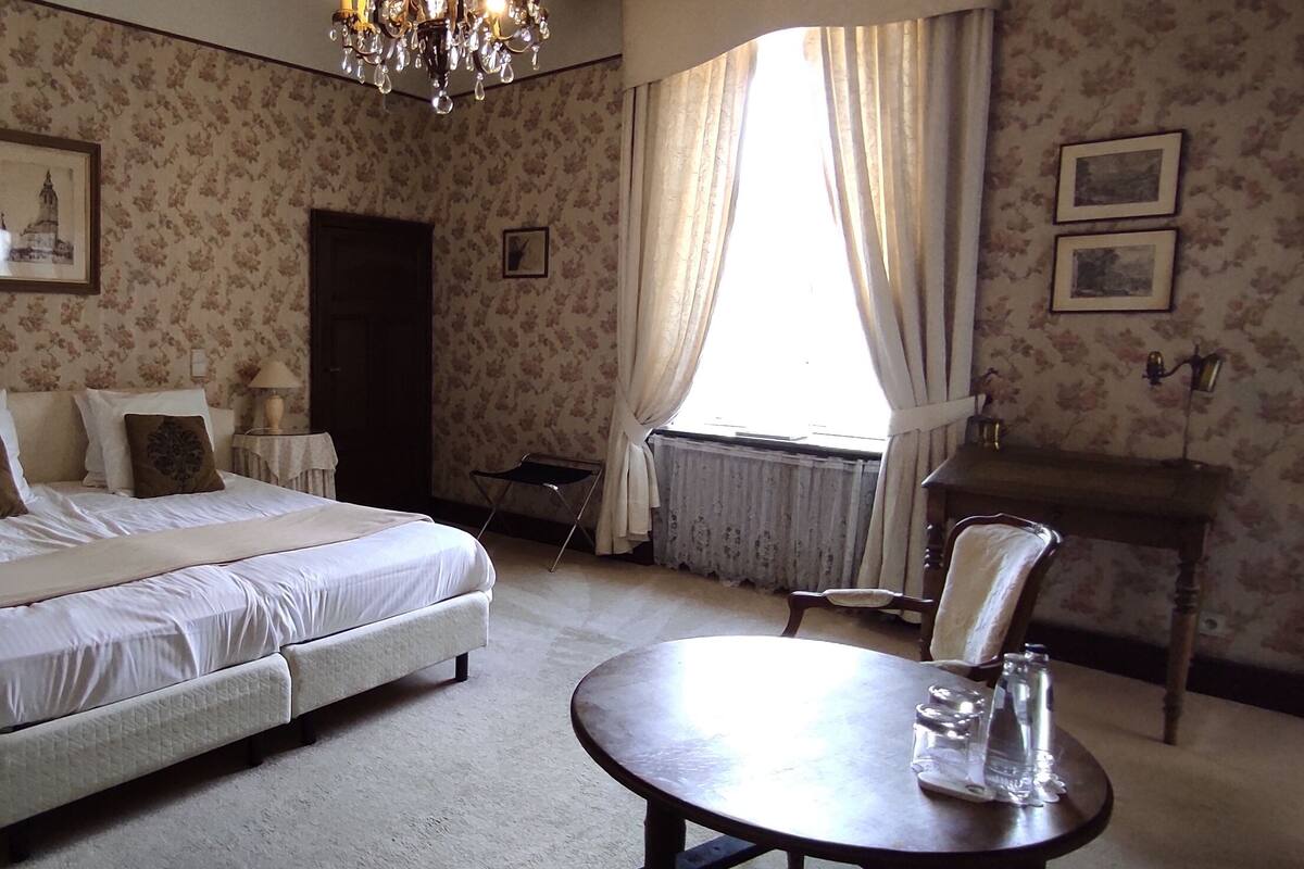 A bedroom with a large bed, a round table, and a chair. There is a window with curtains, a radiator, and a chandelier.