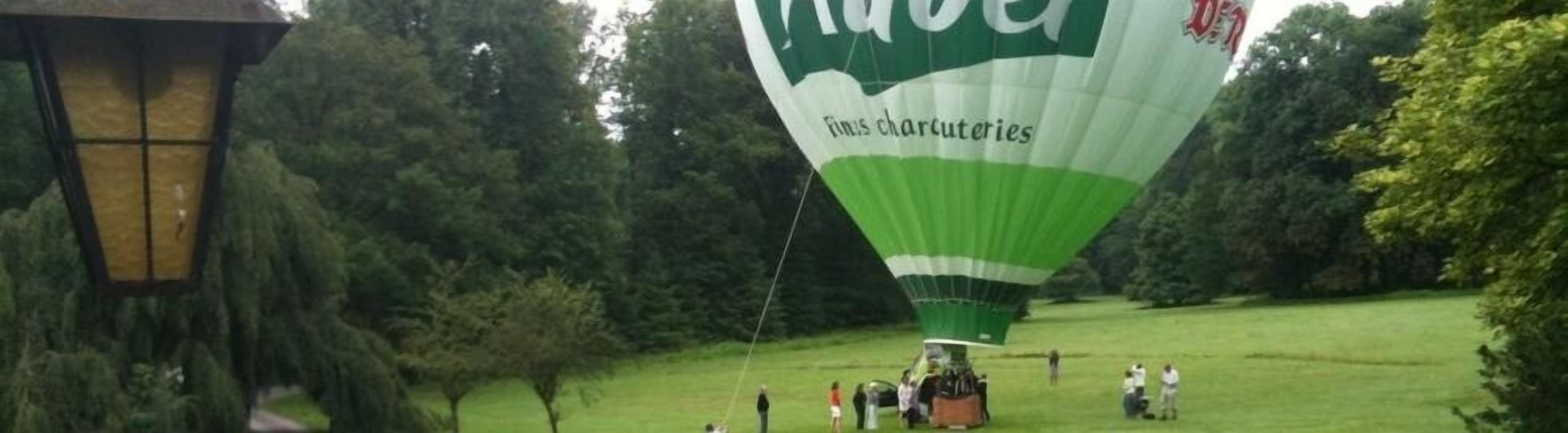 Un globo aerostático con el texto "Fins Charcuteries" preparándose en un campo cubierto de hierba, con árboles al fondo.