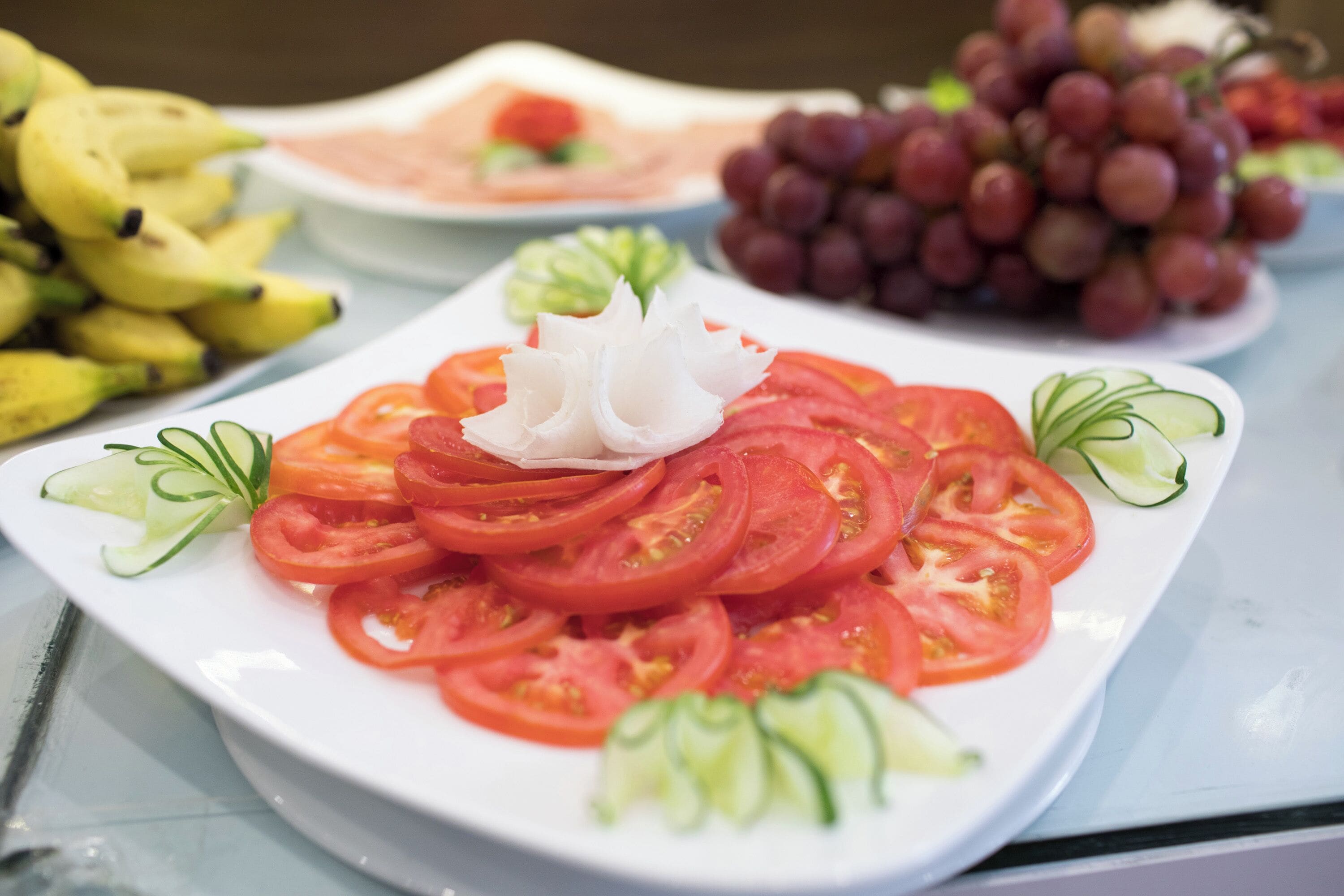 Une assiette de tomates coupées en tranches, agrémentée d’une fleur blanche décorative, entourée de rondelles de concombre et de quelques herbes, avec des bananes et des raisins en arrière-plan.