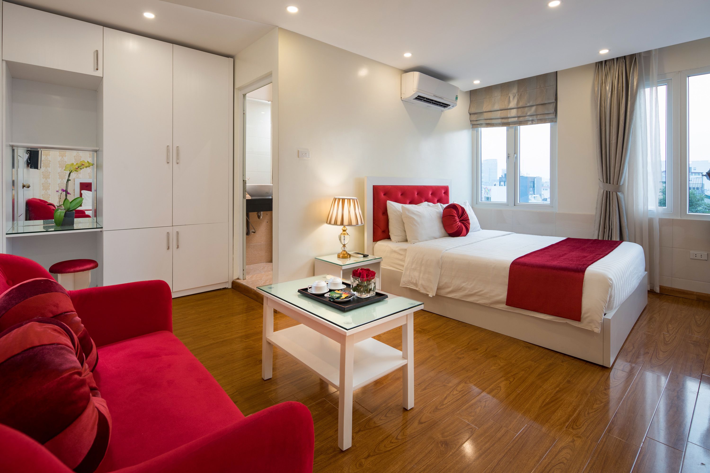 A modern hotel room with a bed, a sitting area with a red chair, a small table with a tray of items, and a wardrobe.