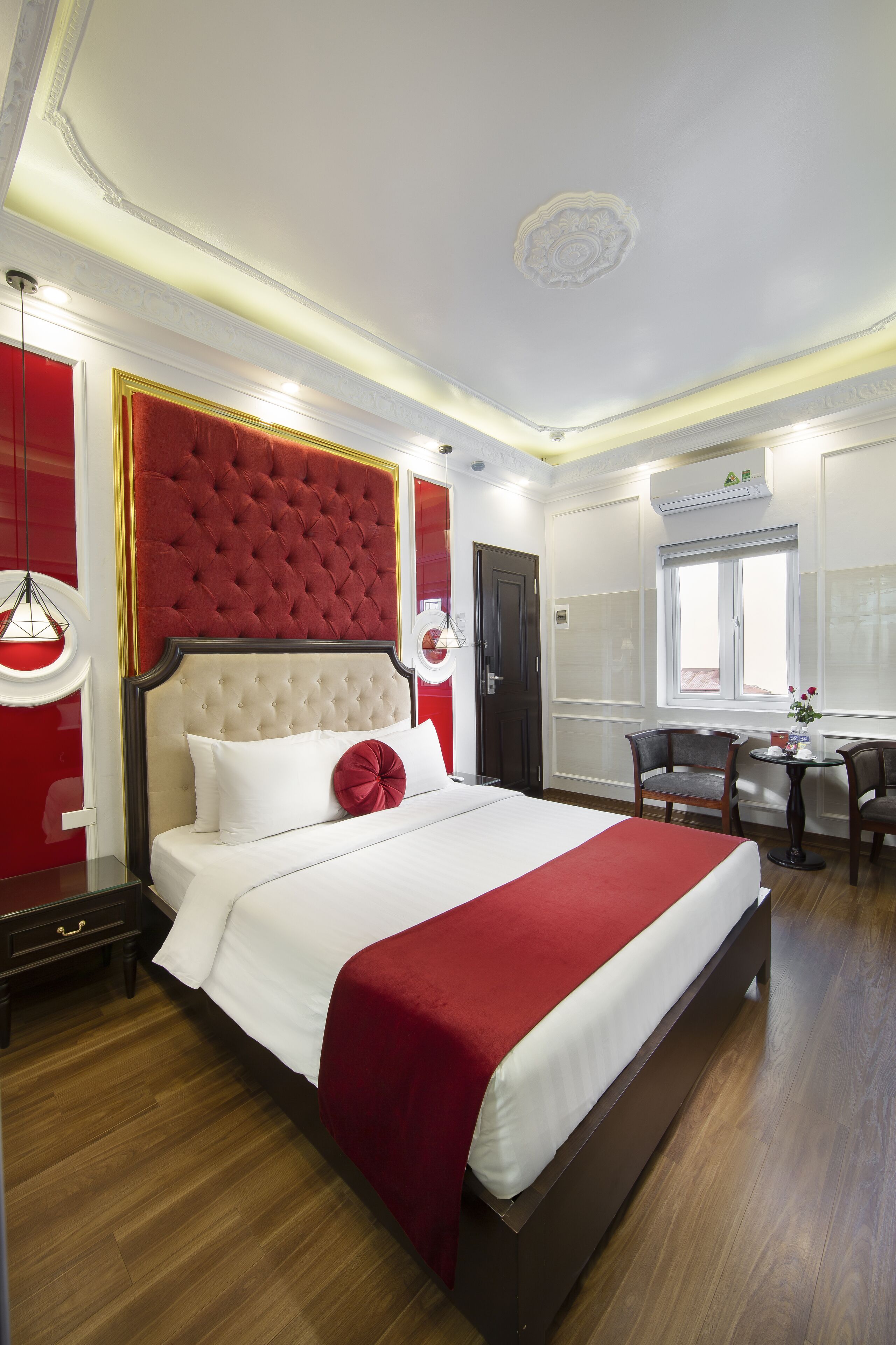 A hotel room with a large bed, a red upholstered headboard, a wooden floor, a desk with two chairs, and a window with a view.