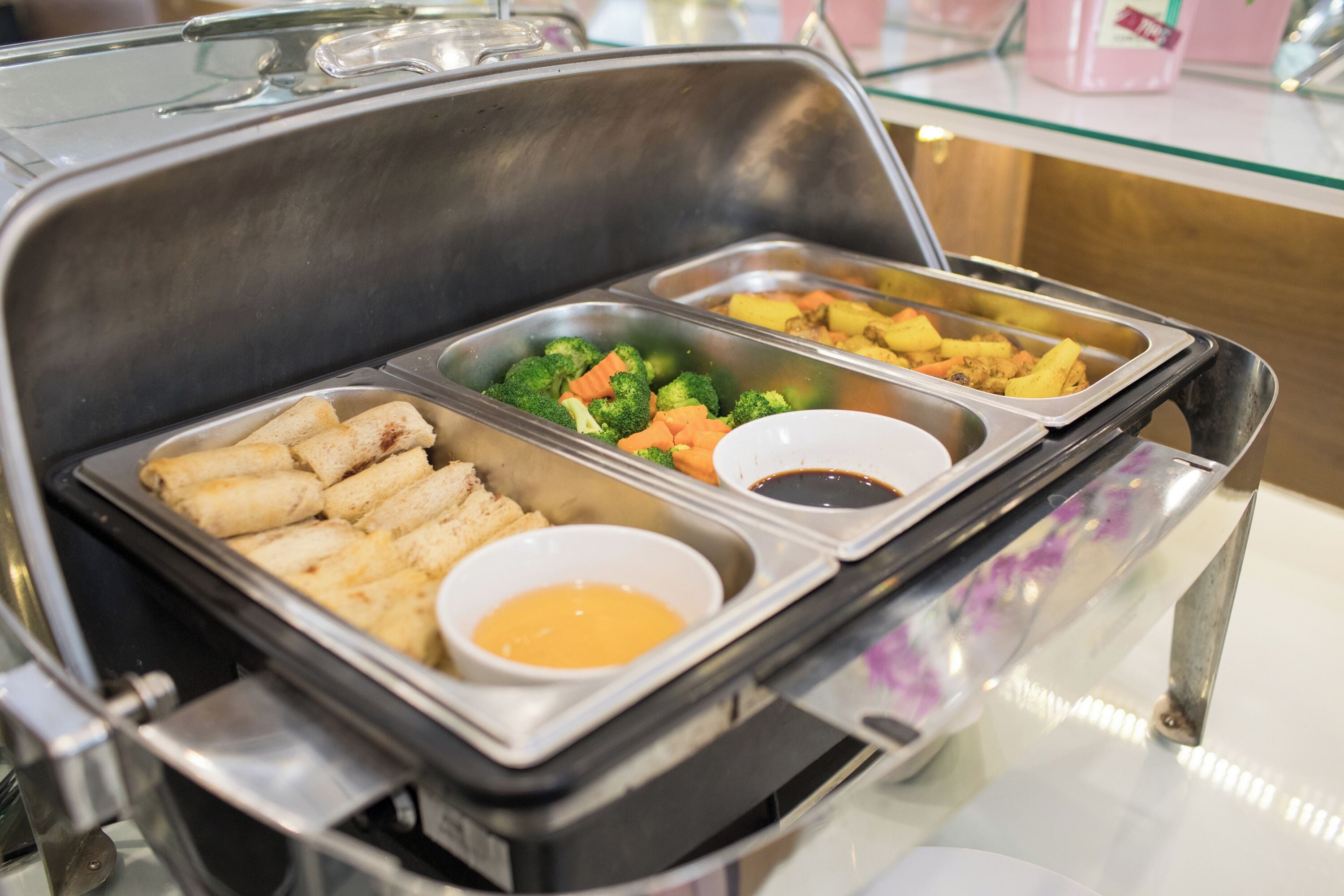 A buffet with various food items including spring rolls, broccoli, carrots, and a sauce container.