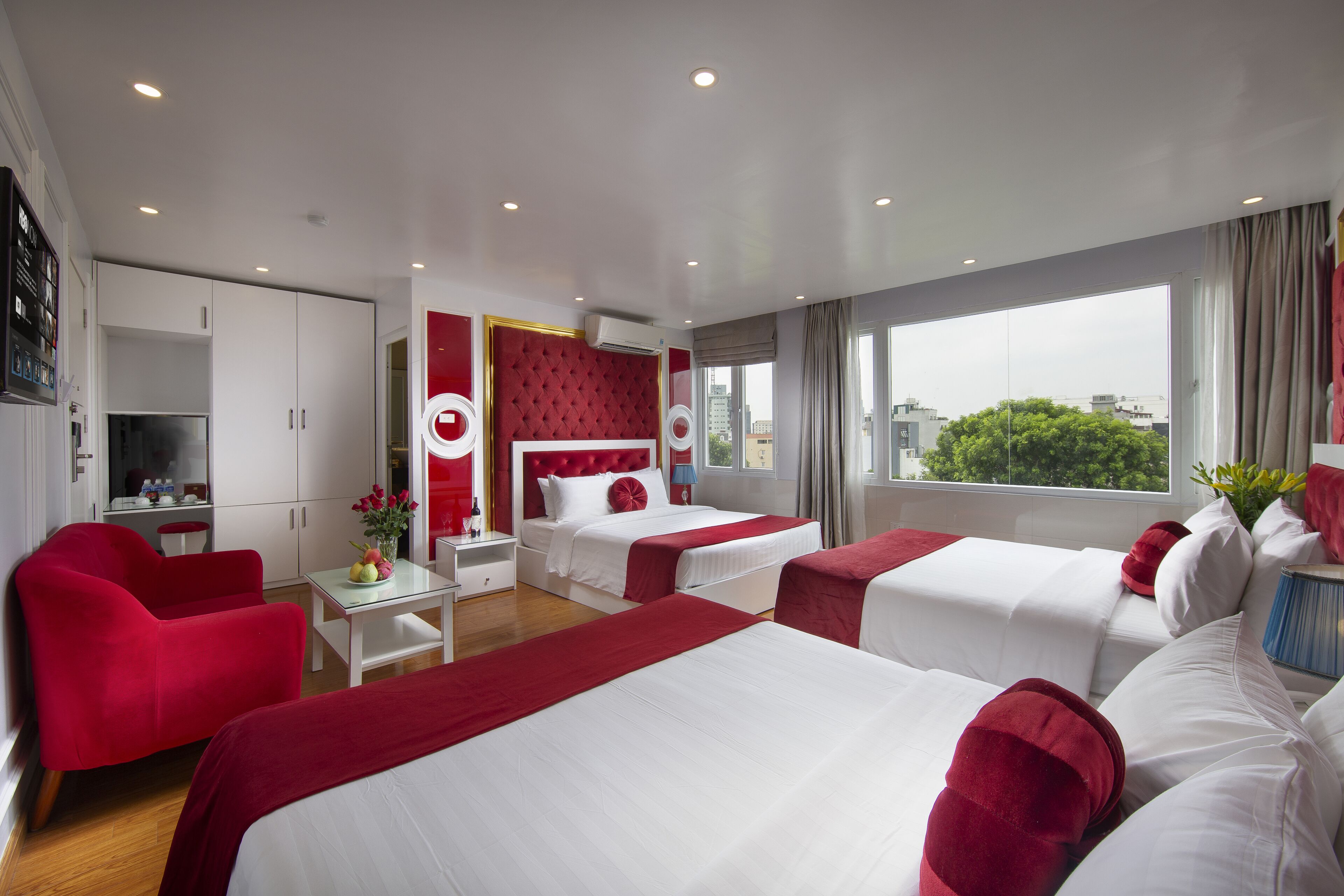 Ein Hotelzimmer mit zwei Betten, einem roten Sessel, einem kleinen Tisch mit Blumenvase und Blick auf die Stadt durch große Fenster.