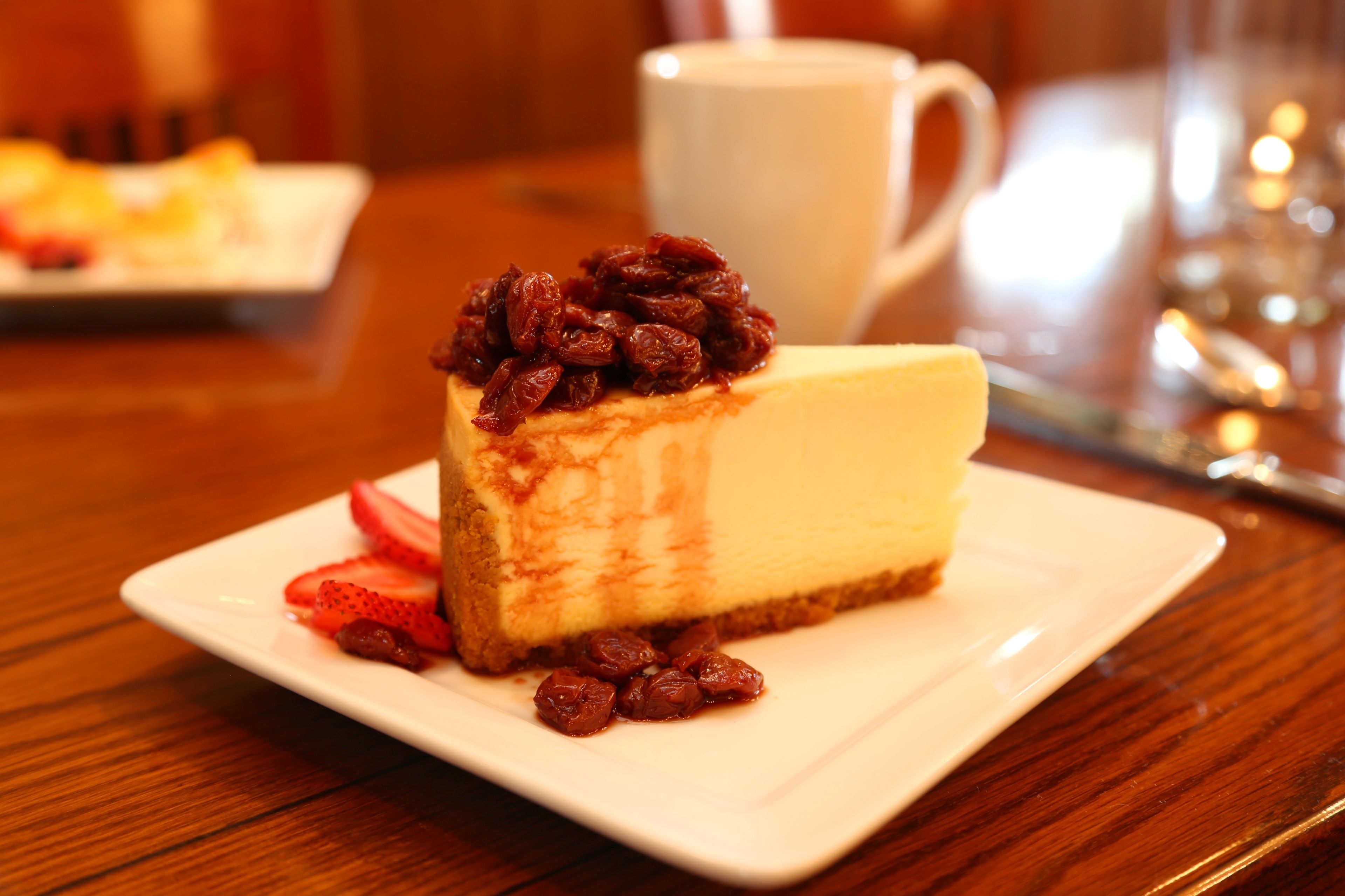 A slice of cheesecake with a fruit topping on a white plate, a white cup in the background, and a wooden table.