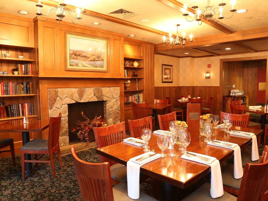 A dining area with wooden tables, chairs, and a fireplace. There are bookshelves with books and decorative items.