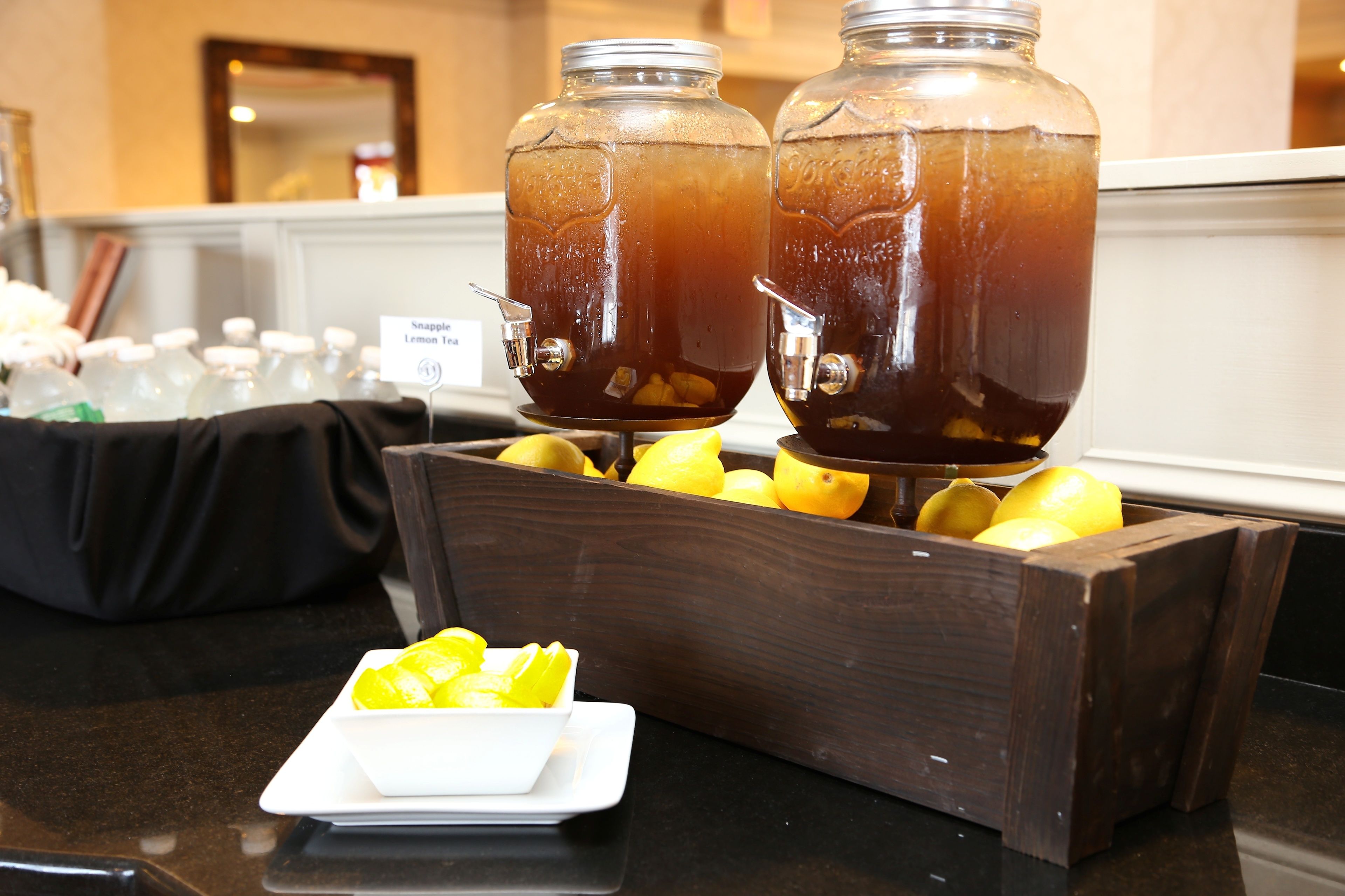Two large glass dispensers filled with iced tea, a wooden crate with lemons, and a small white bowl with lemon slices on a countertop.