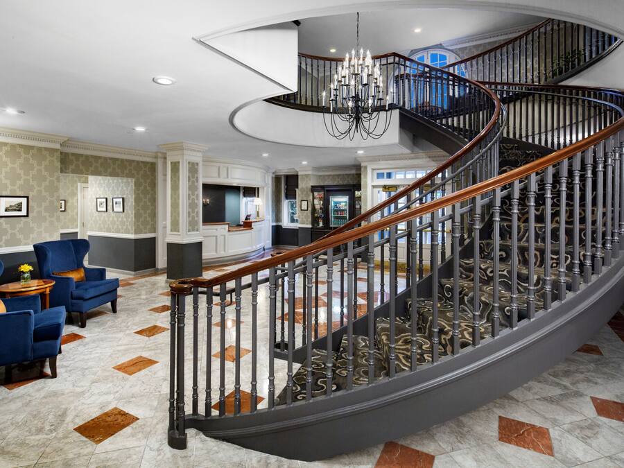A grand spiral staircase with a chandelier, marble flooring, and blue armchairs in a spacious lobby area.